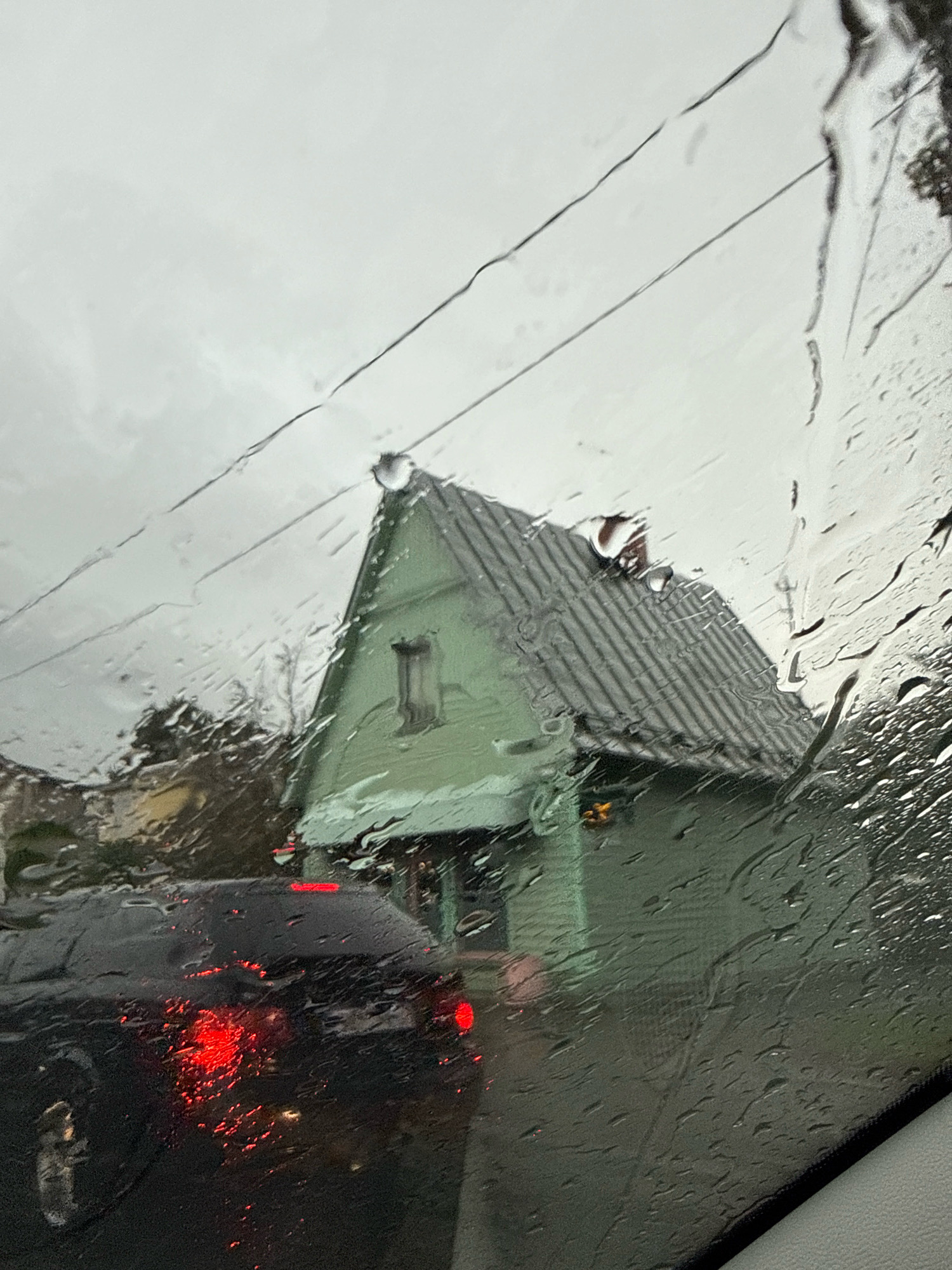 The image, taken from inside a vehicle through a rain-streaked windshield, depicts a light green A-frame house with a dark grey, ridged roof. A single, small window with dark trim is visible on the front gable. Two dark electrical wires cross diagonally in the upper portion of the frame. In the foreground, the rear of a black car is visible, with its red brake lights illuminated. The scene appears to be set on a cloudy, overcast day. There is no discernible information within the image to suggest the location is in Tallinn, Estonia.