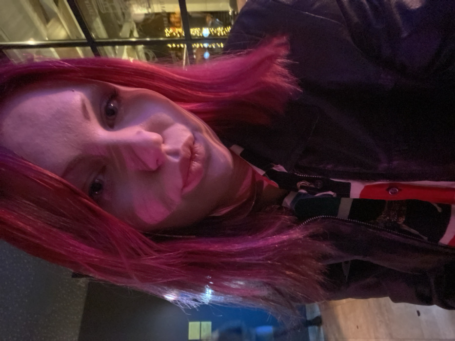 A person with bright pink hair is looking at the camera at an angle. The lighting is low, with a pinkish hue. They are wearing a black leather jacket over a patterned shirt with red, white, and green elements. A window with lights is partially visible in the background, adding to the dim atmosphere.