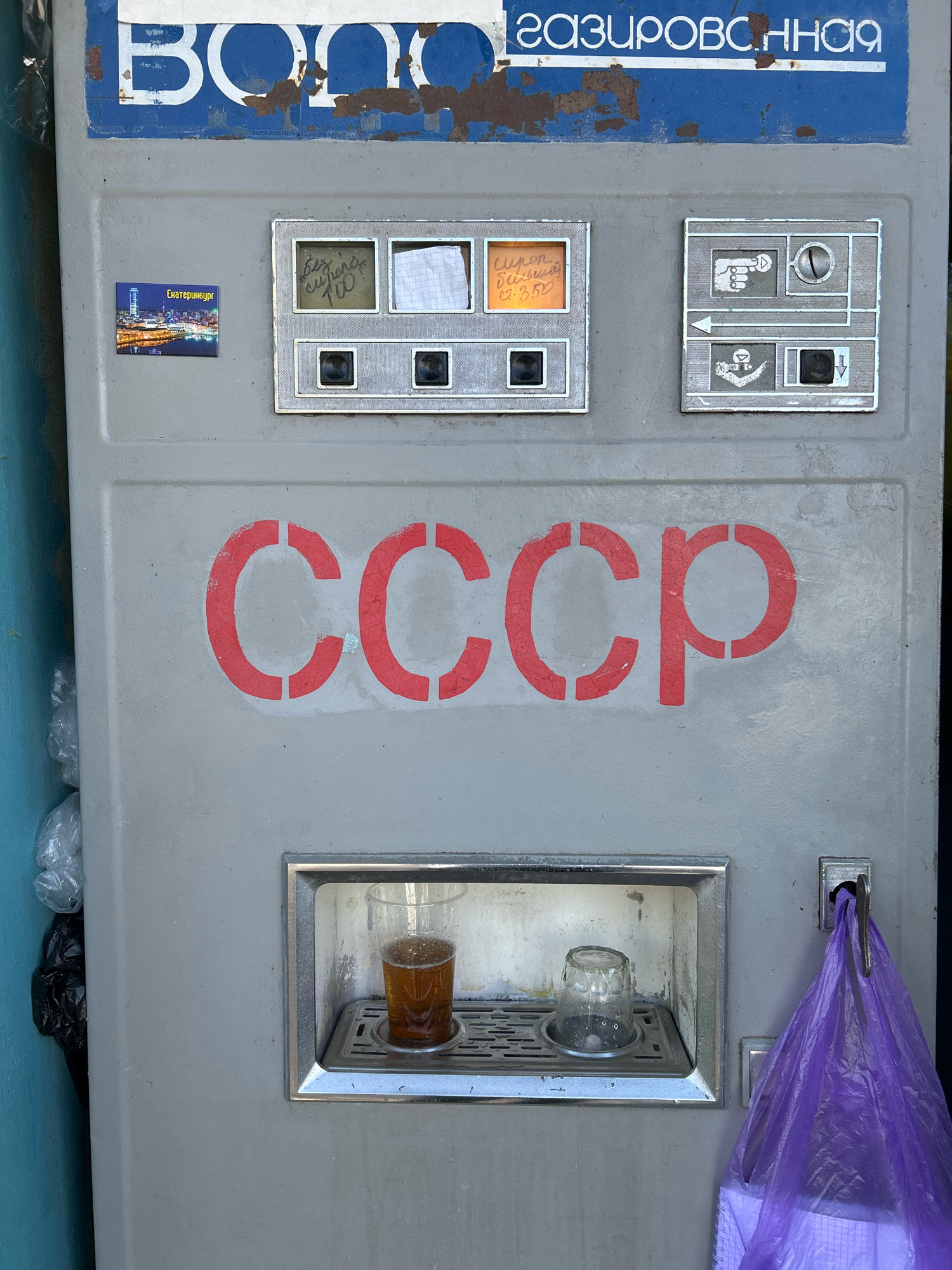 A weathered, gray, Soviet-era carbonated water vending machine stands against a light blue wall. The top of the machine features blue lettering on a white background, partially obscured by rust. Below this, two small rectangular panels are visible. The left panel has a magnetic sticker depicting the skyline of Yekaterinburg, Russia. The panel to its right displays handwritten notes in a small compartment. To the right of these panels is a coin slot and other operational symbols. Dominating the center of the machine's front is the large, red, stenciled word "CCCP" (USSR). Below this, a recessed area holds a clear plastic cup filled with a brown, bubbly liquid, and an empty glass cup. To the right of this dispensing area, a purple plastic bag hangs from a metal hook. The lower section of the machine shows a coin return slot and a button. The overall condition of the machine suggests it is old and has seen significant use, with peeling paint and signs of wear. The presence of the Yekaterinburg sticker on a machine that might be found in Arkalyk, Kazakhstan, suggests a possible connection through tourism or relocation of the machine. The scene conveys a sense of nostalgia for the Soviet era, juxtaposed with the functionality of a public utility.