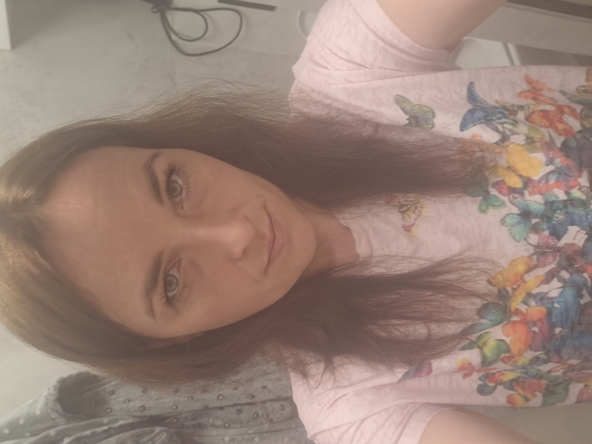 A woman with long, brown hair is taking a selfie in what appears to be a bathroom. She is wearing a light pink t-shirt with a colorful butterfly print. The background shows a tiled wall, a garment hanging on the left, and electrical cords on the right. The overall lighting is soft and diffused. The specific location in Mińsk Mazowiecki, Poland, cannot be determined from the image alone.