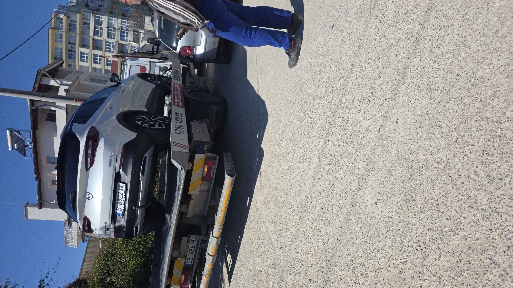 A white Opel Grandland X with license plate B 8922 XK is being towed on a tow truck. The tow truck's license plate is CB 1656 PC. A man wearing blue trousers and sneakers stands near the tow truck. The background shows a building with many windows, and the image appears to be taken in an urban area in Varna, Bulgaria, based on the license plates.
