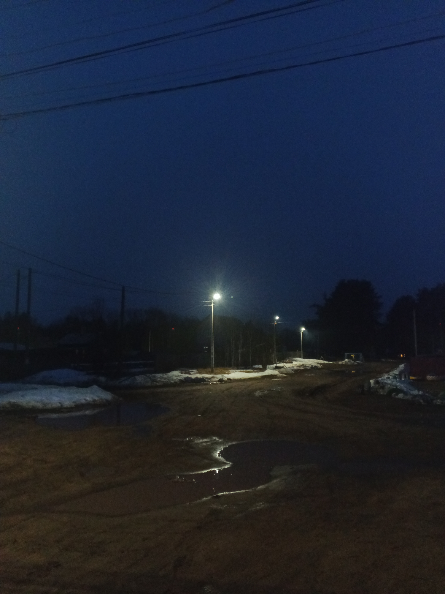 The image depicts an unpaved street in what appears to be the outskirts of Rusalevka, Russia, at dusk or night. Several puddles are visible on the muddy ground. Patches of snow remain on the sides of the road. Streetlights illuminate the scene. In the background, dark silhouettes of trees and buildings are visible against the deep blue sky. Power lines run across the top of the frame. There are no people visible in the image.