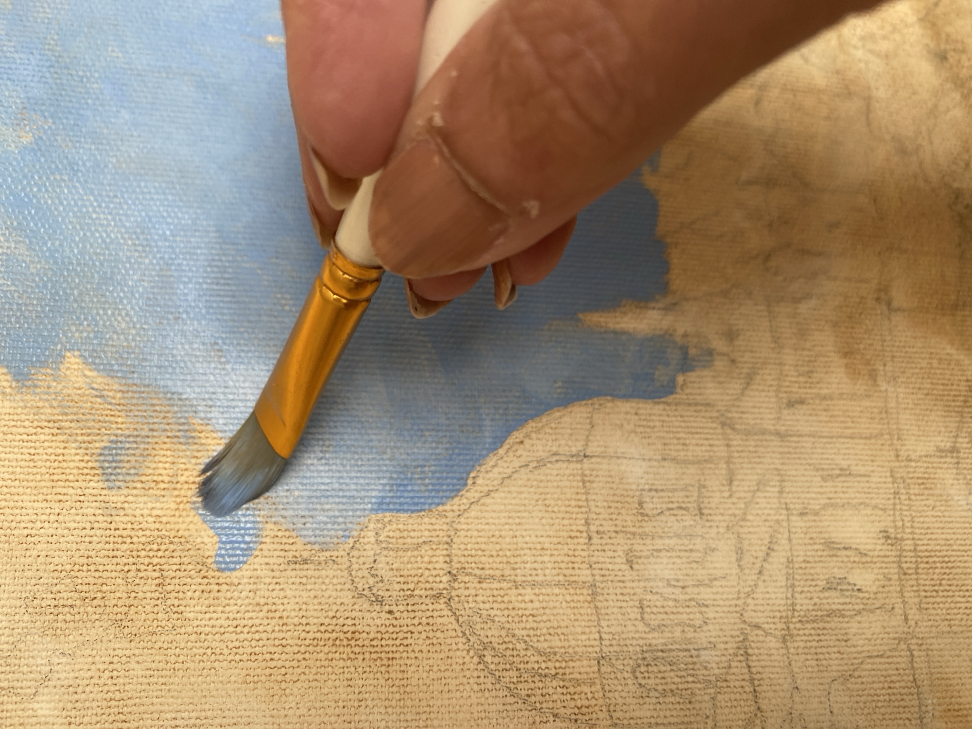 This is a close-up, top-down shot of an artist's hand holding a paintbrush with a golden handle and white bristles. The paintbrush is actively applying blue paint to a canvas with a visible woven texture. The canvas already has faint, preliminary pencil sketches of architectural elements, suggesting a cityscape. A large dome-like structure and other building outlines are discernible in the lower left of the image. The background of the canvas is a light blue color, partially covered by the blue paint being applied. The artist's skin tone is light brown, and their fingernails are painted a similar shade. The scene appears to be an indoor art studio or a similar creative space. The location of Bílina, Czechia, is not visually represented in the provided image.