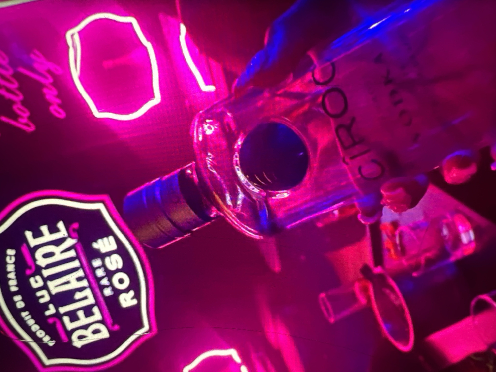 In a dimly lit, vibrant environment illuminated by neon pink and blue lights, a bottle of Cîroc vodka is held by a person whose hand, with painted nails, is visible. The bottle's label, "CIROC VODKA," is partially legible. In the background, a neon sign for "LUX BELAIRE RARE ROSÉ" is prominent, along with another neon sign that reads "bottle only." Several shot glasses and an ice bucket are also visible, suggesting a bar or club setting. The overall impression is one of a lively nightlife scene, possibly in Lagos, Nigeria, given the context provided.
