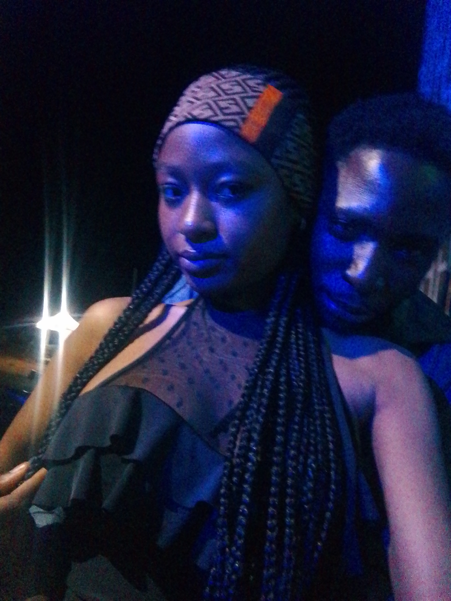 In a dimly lit environment, a young woman with dark braided hair and a patterned headband, accented with an orange stripe, poses for a selfie. She is wearing a black top with a textured yoke and ruffled details. A young man with short hair is positioned behind her, his head resting on her shoulder. The scene is illuminated by blue and purple lighting, casting a faint glow on their faces and skin. The background is dark, with some indistinct lights visible, suggesting an outdoor or dimly lit indoor setting, possibly a club or event.
