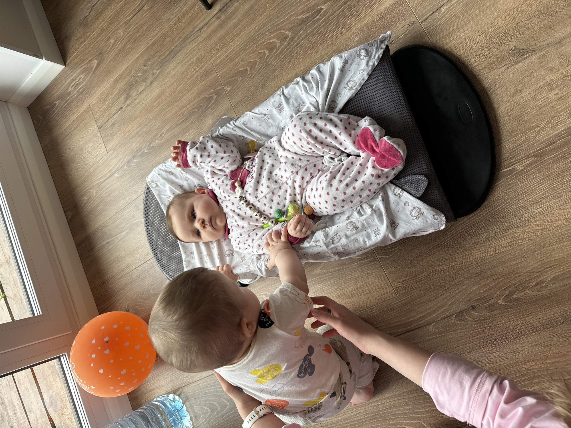 A photograph taken indoors in Chaikovsky, Russia, depicts two infants on a wooden floor. One infant lies on their back in a baby seat dressed in a white pajama with pink hearts and bows, holding a multicolored toy. The other infant is seated on the floor, facing away from the camera, wearing a light-colored pajama with cartoon animal designs and reaching toward the infant in the baby seat. An adult's hands support the seated infant. An orange balloon with white heart designs and a bottle of water are next to the seated infant near a window.