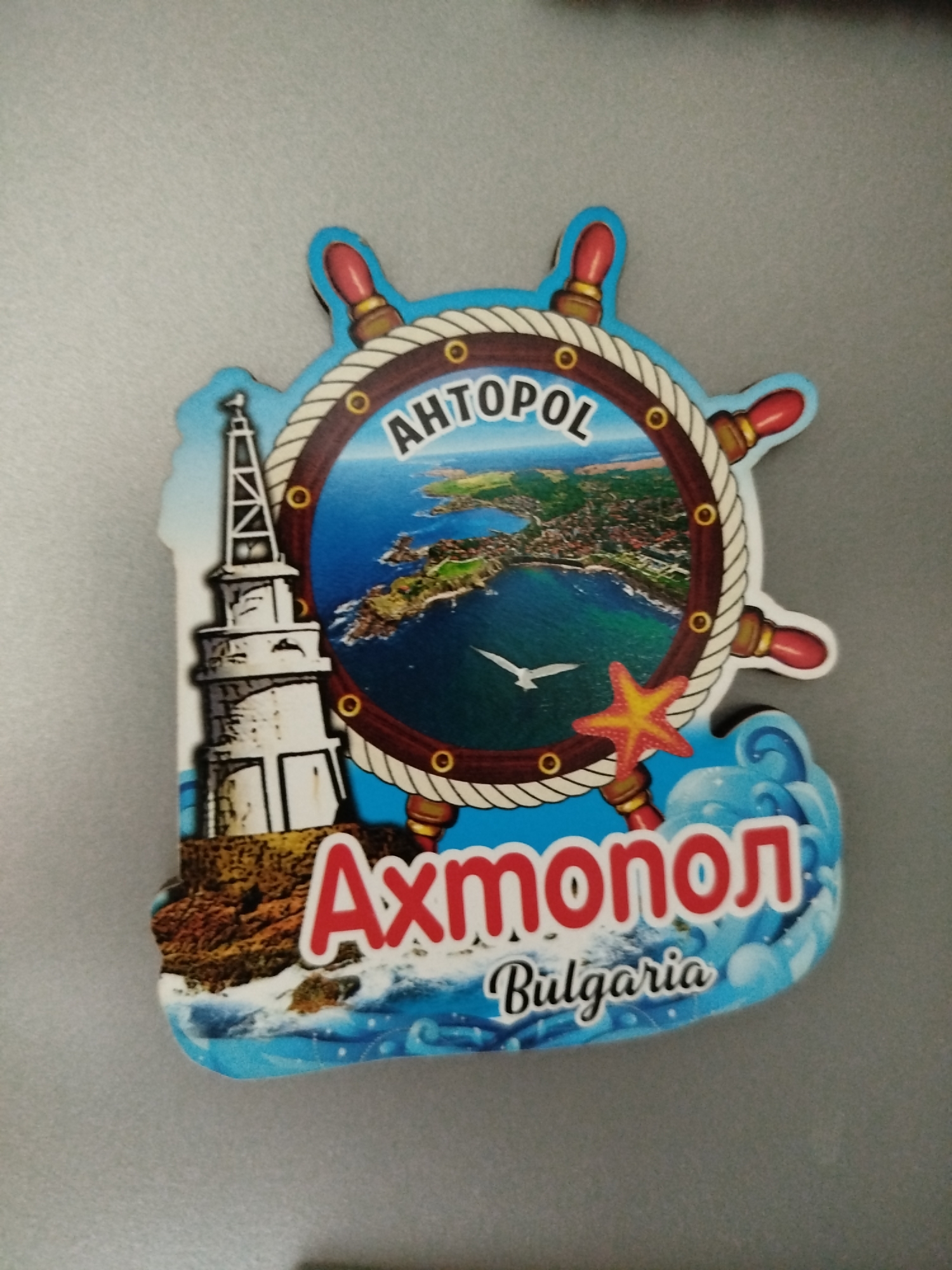 The image shows a magnet souvenir depicting Ahtopol, a town in Bulgaria. The magnet is shaped like a ship's wheel. Within the wheel is a view of Ahtopol's coastline and town. To the left of the wheel is a depiction of a white lighthouse on a rocky outcrop. Below the wheel, in red letters, is the name of the town "Ahtopol", and below that, in smaller letters, is the country "Bulgaria". A starfish is visible on the right side of the wheel. The lower portion is designed with blue waves.