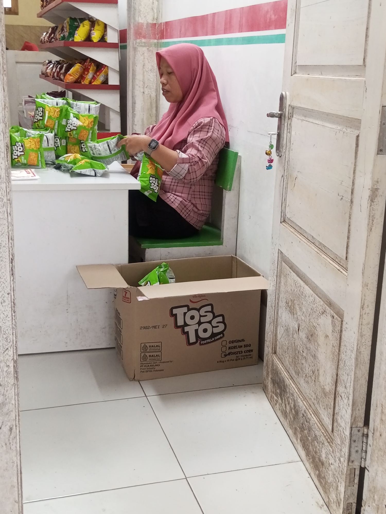 In Kalibeber, Indonesia, a woman wearing a pink hijab and a plaid shirt is seated at a white table. She is actively handling several green bags of chips, suggesting she is either unpacking or organizing them. The table is covered with stacks of these green chip bags, some open and others closed. To her right, a partially opened cardboard box with the "TOS TOS" logo is visible, also containing chip bags. Behind the woman, shelves are stocked with various chip bags. The scene takes place indoors, near a closed white wooden door with a metallic handle. The floor is tiled in a light color.