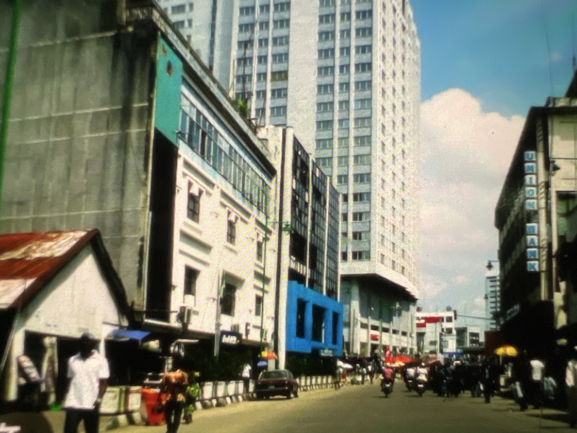 The image depicts a street scene in Lagos, Nigeria, characterized by a blend of modern high-rise buildings and older structures. In the foreground, a street is bustling with activity. Several motorcycles and a few cars are visible on the road, suggesting vehicular traffic. Pedestrians are also present, some walking, others seemingly engaged in commerce or daily routines.
The architecture varies significantly. To the left, a modest white building with a rusty red roof stands. Adjacent to it is a more modern, multi-story building with a striking turquoise facade on its upper section and white lower levels featuring numerous windows. Behind these, a very tall, white high-rise building dominates the skyline, its facade covered with a grid of many small windows, indicative of modern office or residential accommodation. Further down the street, another tall building with a darker facade is visible, featuring a vertical sign with blue letters that spell out "BANK."
The overall impression is of a busy urban environment in Lagos, where contemporary urban development coexists with older, perhaps more traditional, street-level infrastructure. The sky above is partly cloudy, indicating fair weather. The image's quality, with visible digital noise and color aberrations, suggests it was captured with a lower-resolution device or under challenging lighting conditions, possibly a digital photograph with significant compression or artifacting.
