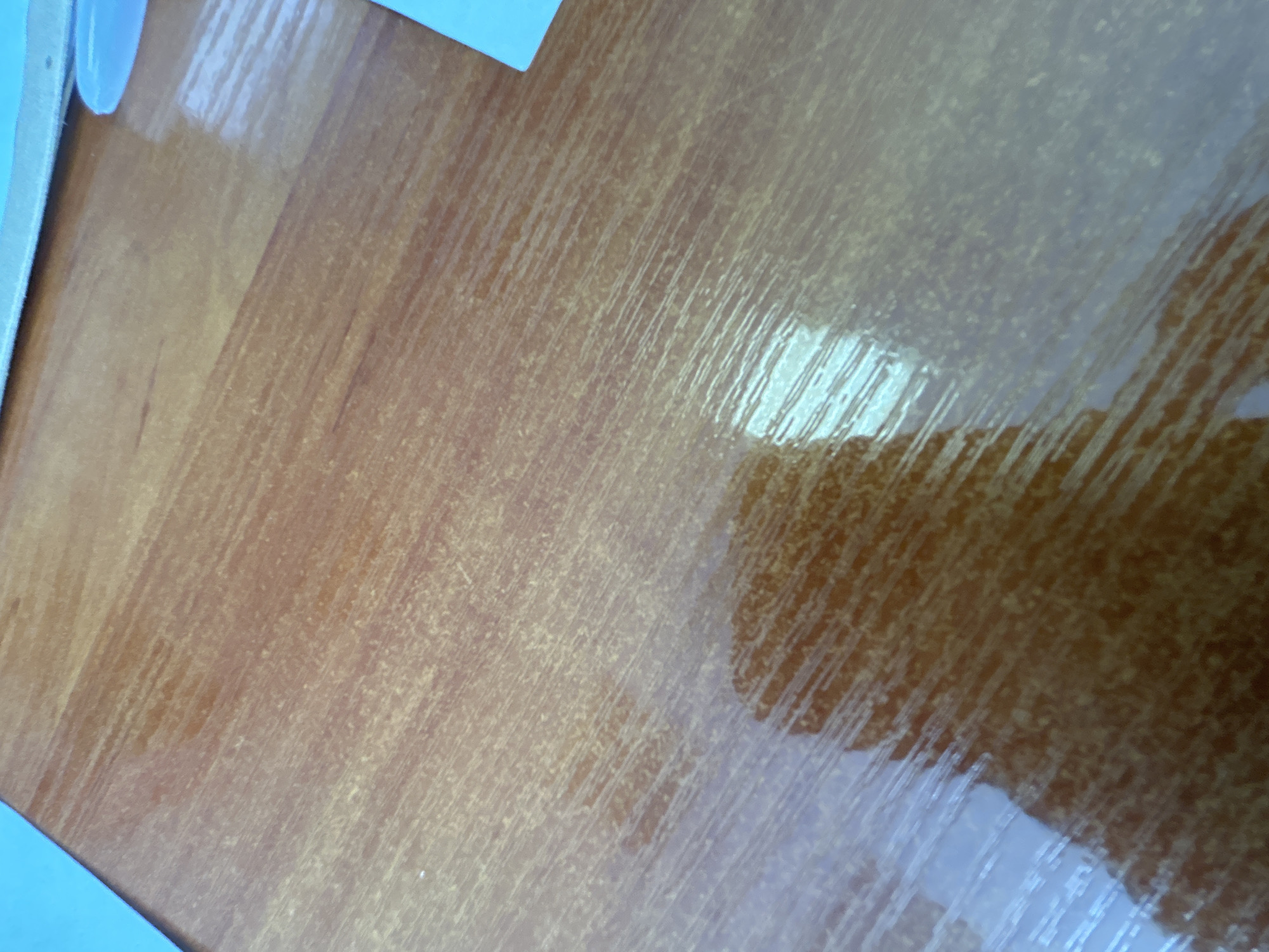 This is a close-up, angled shot of a polished wooden surface, likely a table or desk. The wood has a warm, reddish-brown hue with visible grain lines running diagonally across the frame. The surface is highly reflective, catching light and creating bright streaks and patches that obscure the wood texture in places. The reflections suggest an overhead light source.
In the upper left corner, a small portion of a light-colored, curved object is visible, possibly a mug or a container, with a thin white edge. A sliver of a light blue surface is also present in the far left and lower left corners, suggesting it's a larger object or the edge of the table itself.
The image is an abstract study of light, texture, and color on a mundane surface. There are no people, and no specific activity or event is depicted. The lighting creates a dynamic interplay of light and shadow, highlighting the polished finish of the wood. The overall impression is one of an indoor environment with adequate lighting. No text is visible in the image.