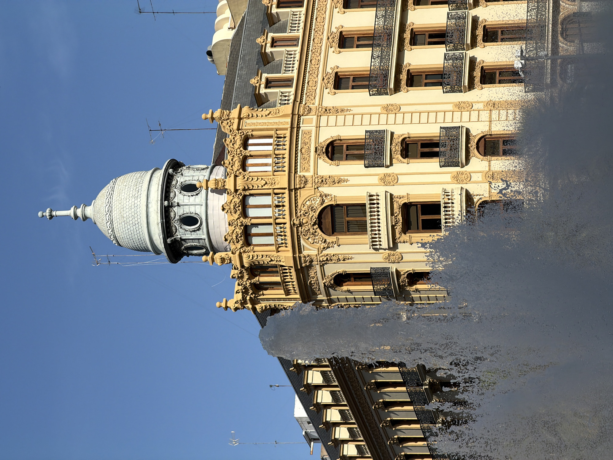 An ornate building with a dome and balconies stands against a clear blue sky, partially obscured by a large fountain in the foreground. The building is cream-colored with detailed yellow ornamentation around its windows and along its cornices. Several floors are visible, each featuring numerous windows, some with decorative iron balconies. The dome, positioned at the top of the building, has a rounded shape and is capped with a finial. Antennas are visible on the roof. The fountain sprays a large volume of water upwards and to the right, creating a dynamic, misty effect that partially obscures the lower and central portions of the building. The scene is illuminated by bright sunlight, casting shadows and highlighting the architectural details.