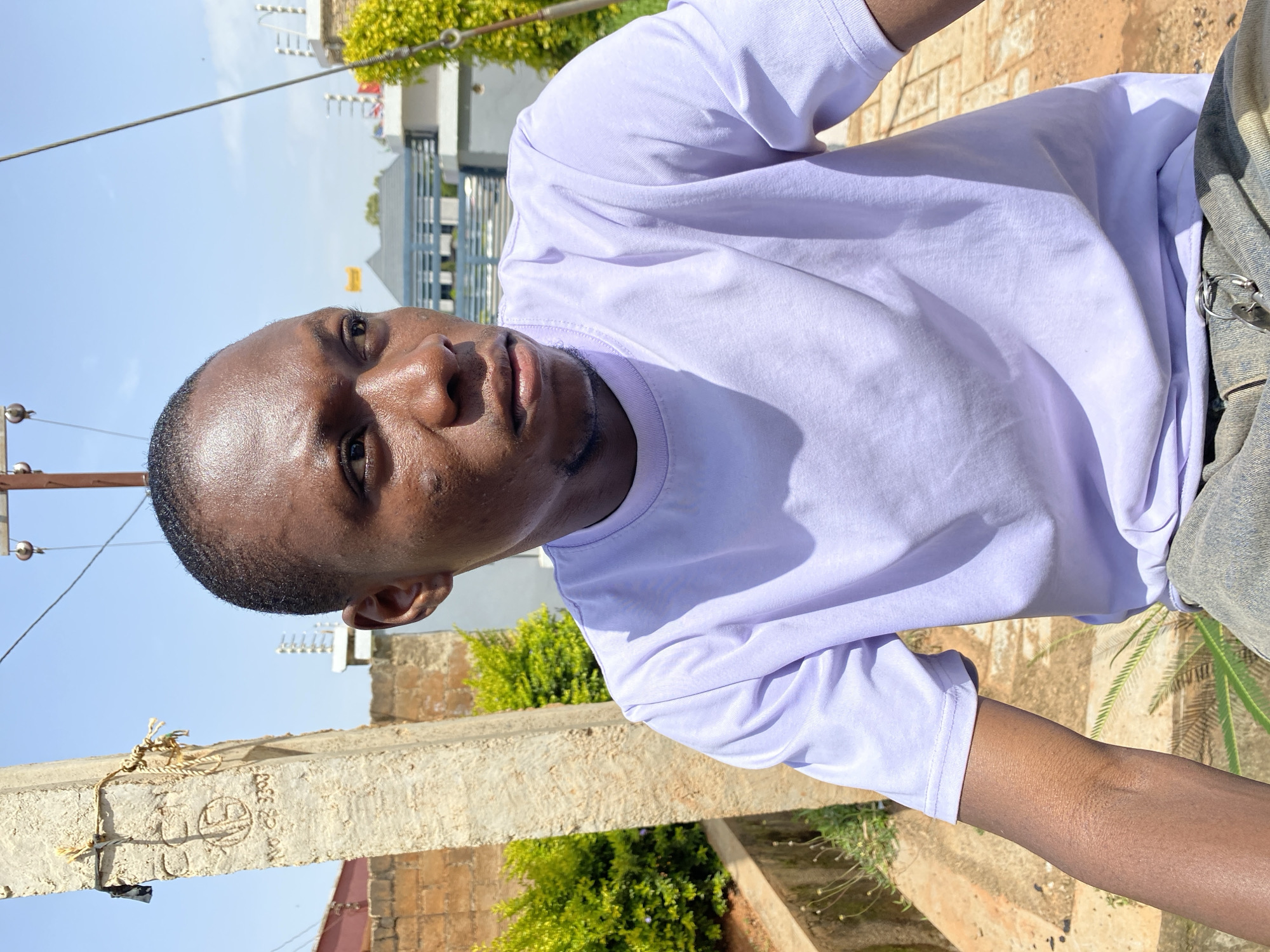 A medium shot shows a young Black man in a light purple t-shirt and blue jeans outdoors. He is looking to his right, with his head slightly tilted. The man has short hair and a light beard. He is positioned in front of a concrete pole with some markings and rope attached. Behind him, there are green bushes, a brick wall, and modern buildings with grey siding and railings under a bright blue sky. The ground is dirt and gravel. The image appears to have been taken during the day in Bukuru, Nigeria.