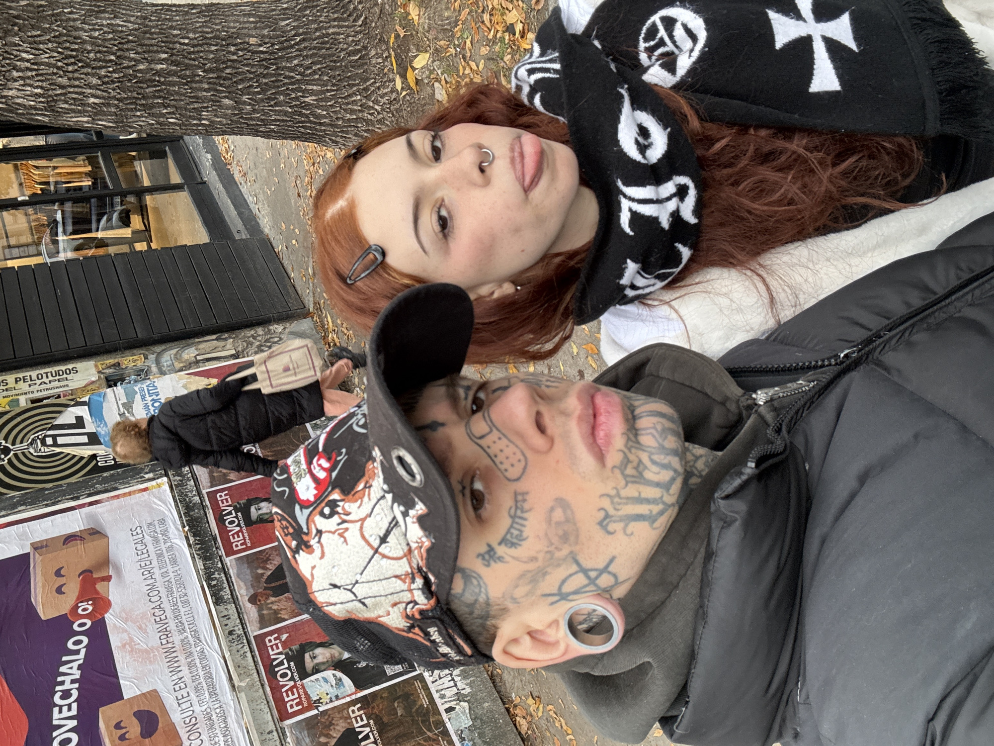 Two people are posing in a photo on the sidewalk in the city of Buenos Aires, Argentina.
The person in the foreground wears a black cap with colorful designs and has tattoos covering their face and a large ear gauge. They are wearing a dark hooded jacket.
Behind them is a person with red hair and a nose ring, wearing a black and white patterned scarf and a white top.
The background shows posters attached to a wall or fence. The posters are advertising "REVOLVER" clothing and a shop called "FRAVEGA". The image also includes glimpses of a shop window and the trunk of a tree. Fallen leaves are scattered on the sidewalk.
