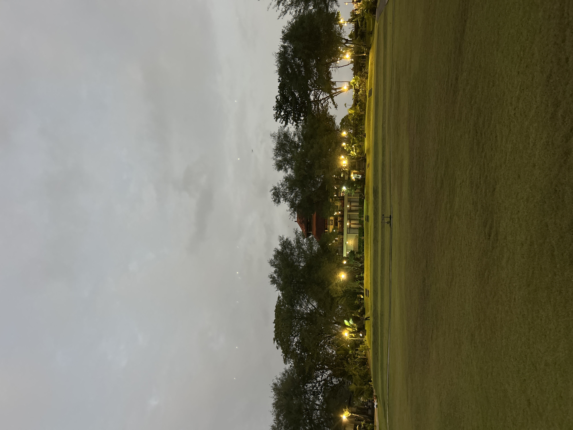 The image depicts a wide, manicured green lawn in the foreground, sloping upwards towards a line of trees. Behind the trees, a building with a red tiled roof and illuminated windows is visible. Numerous yellow lights are strategically placed along the pathway and among the trees, illuminating the area as dusk settles. The sky is overcast and a muted gray. A single person is walking on the lawn in the mid-ground, and a few other indistinct figures can be seen in the distance near the building. The scene appears to be a park or a resort area. The location is identified as the Simpang Lima Gumul (SLG) park in Kediri, Indonesia.
