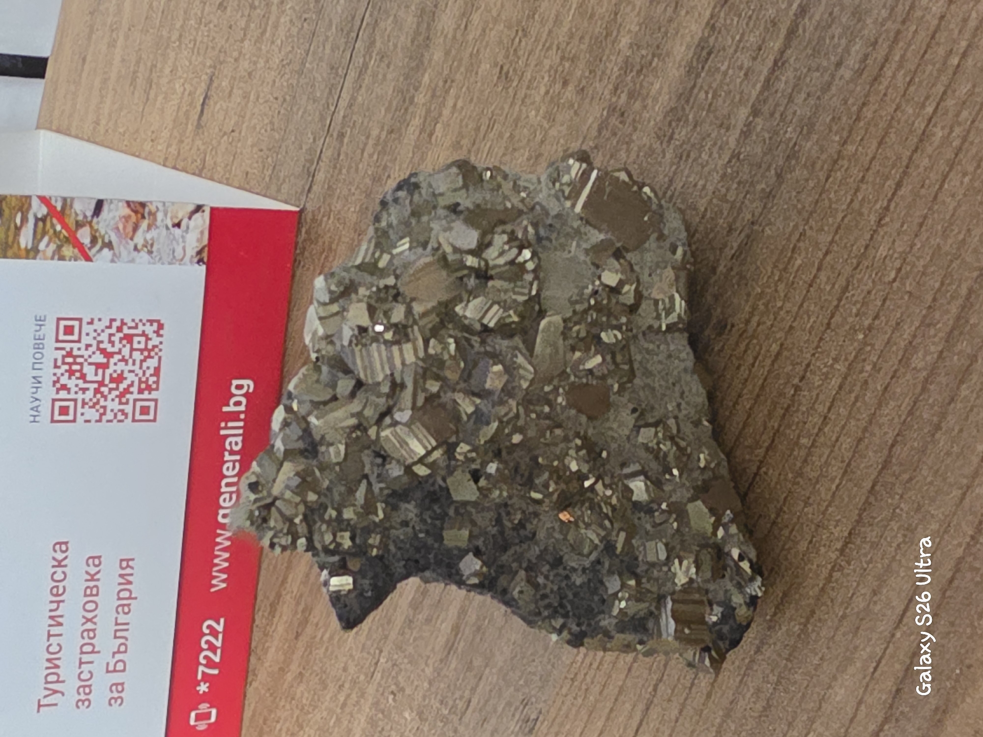 The image displays a piece of pyrite, commonly known as "fool's gold," resting on a wooden surface. The pyrite specimen is multifaceted, with distinct cubic crystals embedded in a darker matrix. Some crystals exhibit a metallic luster and a brass-yellow hue, characteristic of pyrite.
Adjacent to the pyrite is a white and red card. The card has text in Bulgarian, which includes "Туристическа застраховка за България" (Travel insurance for Bulgaria) and "*7222 www.generali.bg". A QR code is also visible on the card. The overall lighting suggests an indoor setting, possibly a desk or table. The wooden surface has a visible grain pattern, indicating it's likely a natural wood material. There are no people or animals in the image, and no specific weather conditions are implied. The scene is static, focusing on the mineral specimen and the informational card. The text on the card, along with the implied context of travel insurance, hints at a commercial or informational purpose for the card's presence. The presence of the "Galaxy S26 Ultra" text in the bottom right corner indicates that the image was captured using a Samsung Galaxy S26 Ultra phone. The location context provided is Plovdiv, Bulgaria.