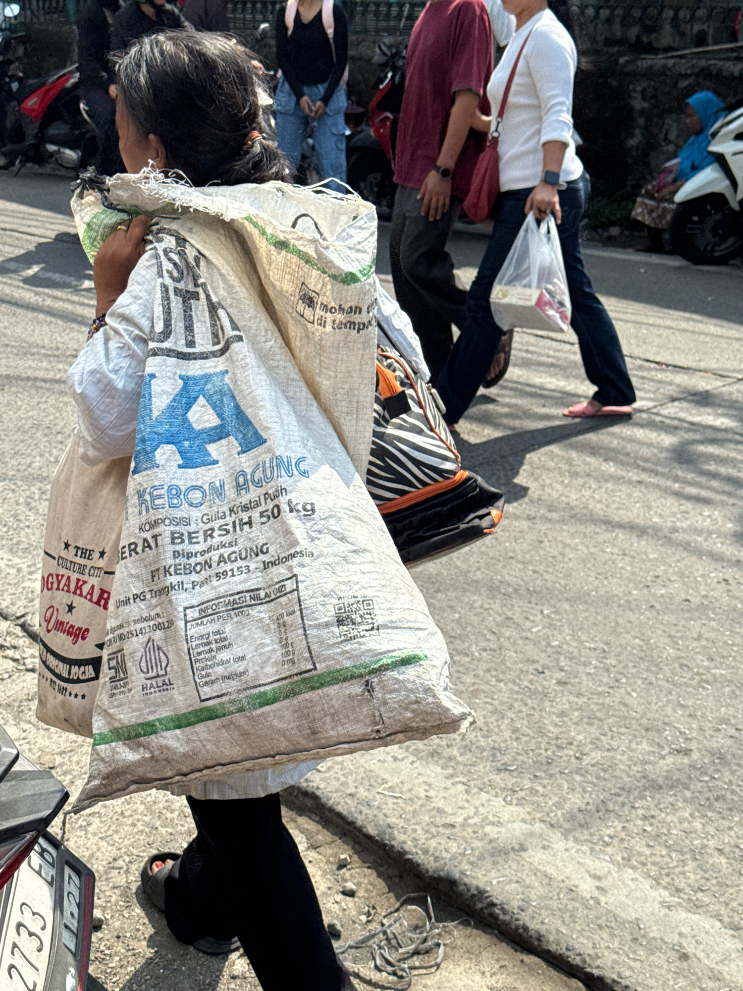 In Sukaraja, Indonesia, a woman carries a large, woven sack of what appears to be sugar on her right shoulder. The sack is white with green and blue lettering, including "KEBON AGUNG", "GULA KRISTAL PUTIH", and "BERAT BERSIH 50 kg." She is wearing black leggings and dark sandals. Behind her, a person in a maroon shirt walks to the left, and another person in a white long-sleeved shirt and blue jeans walks to the right, carrying a plastic bag. In the background, a white motorcycle with blue accents is parked, and a woman in a blue headscarf sits near it. The scene takes place on a paved road with some debris.