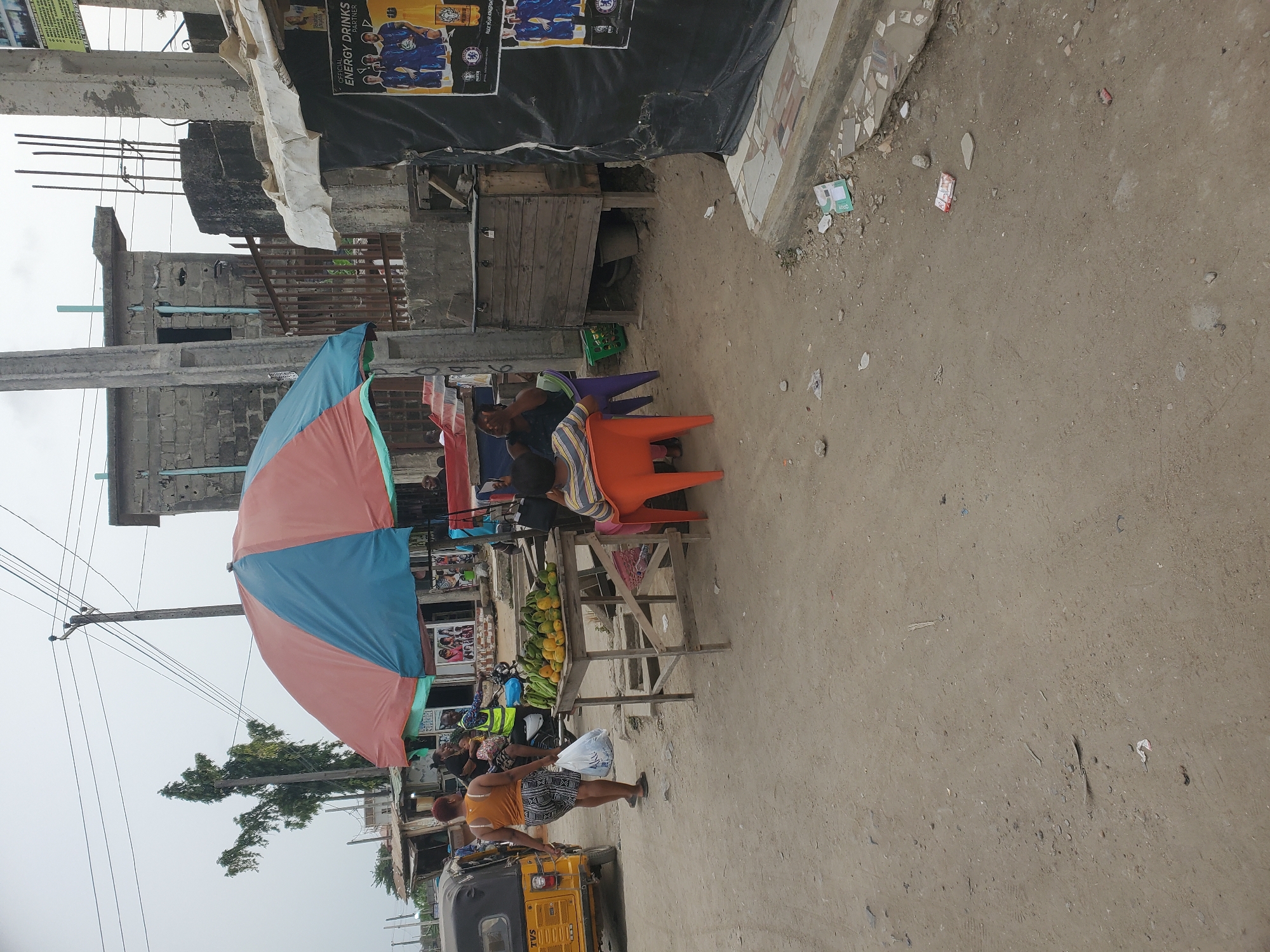 The image depicts a bustling outdoor street market scene in what appears to be Lagos, Nigeria, under an overcast daytime sky. The ground is an unpaved expanse of dirt and gravel, littered with small pieces of debris and discarded wrappers.
In the midground, a large, multi-colored umbrella, featuring segments of blue, red/pink, and light green, offers shade to several individuals. Beneath it, people are seated on plastic chairs of various colors, including bright orange and purple, seemingly engaged in conversation or waiting. To the right of the umbrella, a wooden stall displays a variety of fresh produce, notably bunches of green bananas and yellow fruits resembling oranges or mangoes.
In the foreground, a woman wearing a yellow top and patterned shorts walks to the left, carrying a white plastic bag. A yellow three-wheeled auto-rickshaw, commonly known as a Keke Napep, is partially visible on the far left of the frame. Utility poles with numerous crisscrossing electrical wires are prominent in the upper left, suggesting the local infrastructure.
The background is filled with various concrete block buildings, some appearing unfinished with exposed rebar, a common sight in urban construction in the region. High on a wall above the market area, a black banner or poster is visible. Text on this poster reads "OFFICIAL ENERGY DRINKS PARTNER" and features images of footballers in blue jerseys, alongside logos, one of which appears to be "HYDRATE". The brand "TVS" is partially visible on the yellow auto-rickshaw, and discarded packaging, possibly for "Milo" and another green product, can be seen on the ground. The scene portrays everyday life in a vibrant, functional street market.