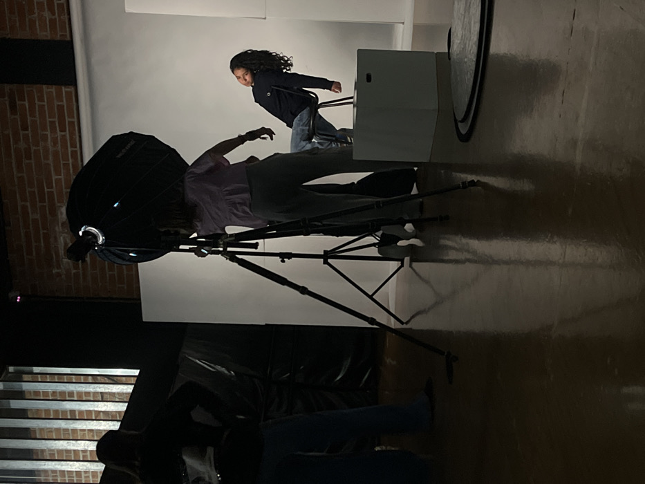 The scene depicts an indoor photography studio setup in Puebla, Mexico. Three individuals are present. In the right midground, a young person with long, dark, curly hair, wearing a dark long-sleeved shirt and blue jeans, is seated on a black chair positioned atop a white cubic block. This individual is angled slightly to their left, leaning back with a slight smile. Immediately to the left of the seated subject, a second individual stands, partially obscured by a tall, black tripod light stand equipped with a large, black parabolic softbox containing a visible light source. This individual, wearing a light-colored top and dark pants, appears to be adjusting the lighting equipment. A third individual is visible on the far left, primarily in silhouette, with hands on hips, facing away from the main setup. The immediate background behind the seated subject and lighting equipment is a seamless white backdrop. To the left, a darker section reveals a brick wall texture with vertical metal bars and a dark, possibly draped, object. The floor is a highly reflective, polished dark surface, showing reflections of the studio equipment. A silver circular photography reflector lies flat on the floor in the bottom right foreground. Artificial studio lighting illuminates the primary subjects and backdrop.
