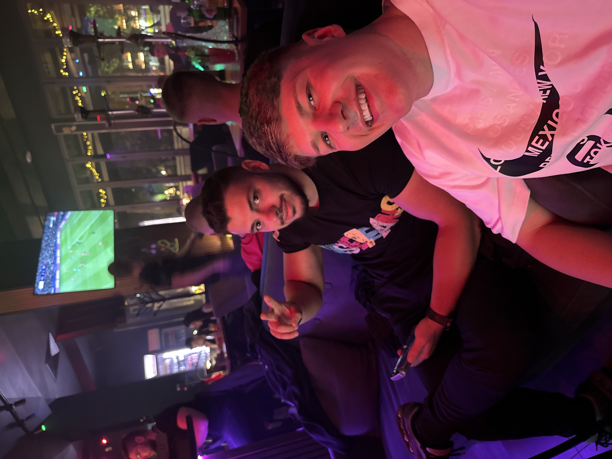 Two young men are captured in a vibrant indoor setting in Plovdiv, Bulgaria, likely a lounge or shisha bar, at night. The image is taken at a Dutch angle, showing a dynamic, social scene.
In the foreground, a young man with short brown hair is smiling broadly, looking directly at the camera. He wears a white t-shirt featuring a large black Nike swoosh logo. Overlapping the logo and shirt are names of cities in a repeating pattern, including "PARIS," "LONDON," "LOS ANGELES," "MEXICO," "NEW YORK," "TOKYO," and "BERLIN." His face is illuminated with a strong red-pink light.
Behind him and slightly to his left, another young man with a beard and dark hair is visible, also looking towards the camera with a slight smile, making a peace sign with his right hand. He is wearing a black t-shirt with colorful graphics and holds a dark game controller in his left hand.
The background is dimly lit with atmospheric red, pink, and purple hues. Through large windows, decorative string lights can be seen outdoors, suggesting an evening or nighttime setting. Further in the background, a television screen displays what appears to be a football or soccer match. Shisha pipes are also visible on tables in the distance, confirming the lounge-like ambiance. Other blurry figures suggest the presence of more patrons, contributing to the lively and relaxed atmosphere. The overall mood is cheerful and social.