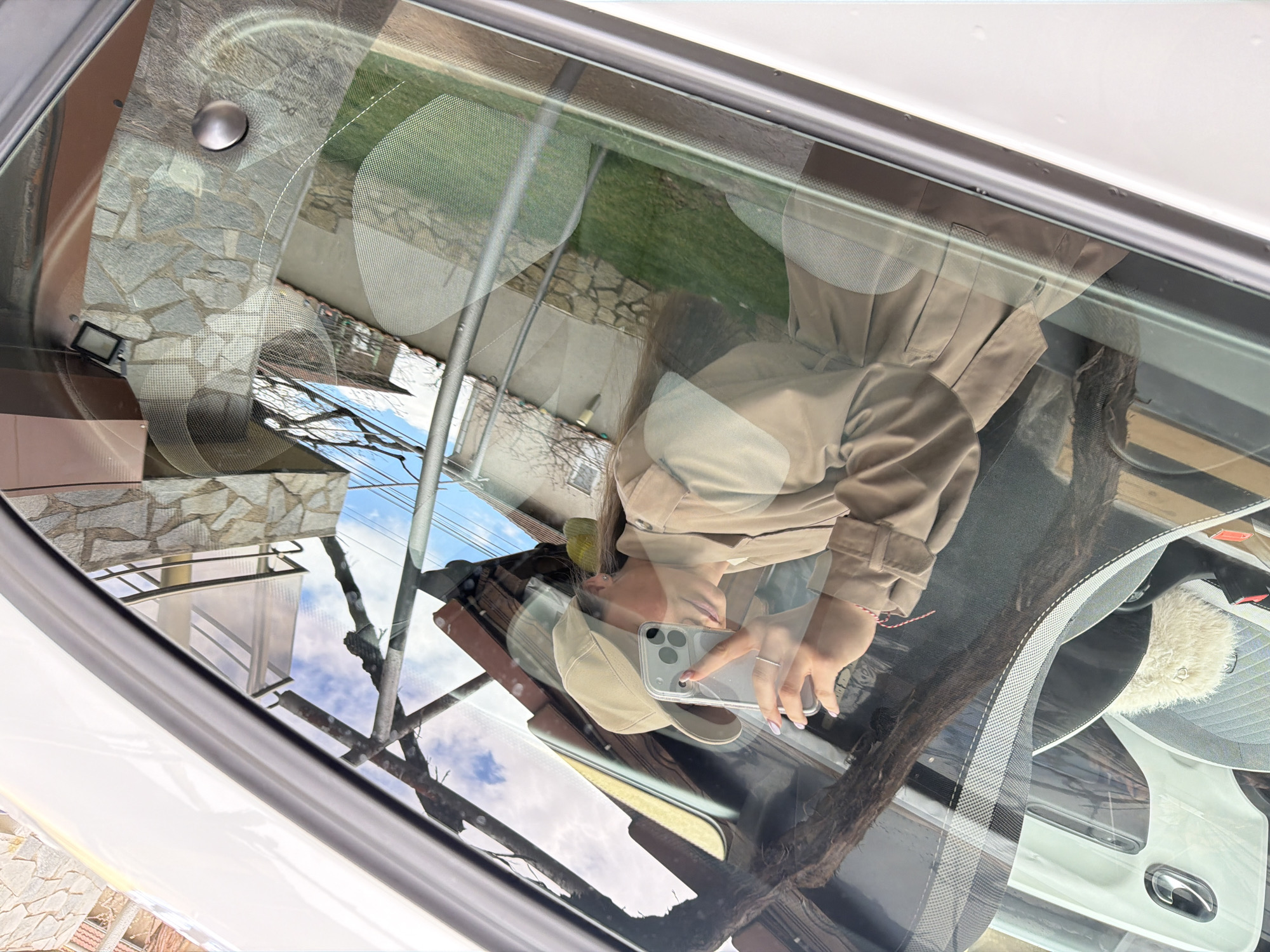 A reflection in a car window shows a person taking a selfie with their phone. The reflection also reveals the exterior environment, including parts of a building made with stone bricks, a grassy area, tree branches against a blue sky, and what appears to be metal support beams. The interior of the car is partially visible, including the dashboard, part of the driver's seat, and the inside of the car door.
Based on the location data, the image was taken in Buhovo, Bulgaria.