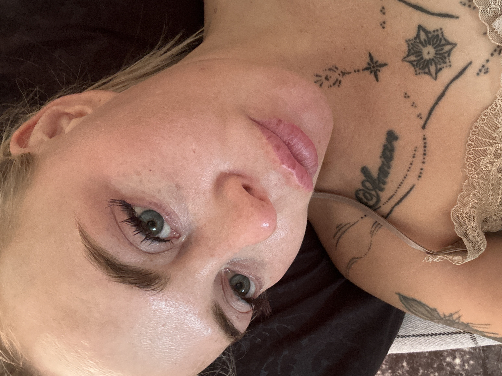 The image is a close-up, high-angle shot of a woman's face and upper chest, taken indoors. The woman has fair skin, light-colored hair, and green eyes. Her makeup includes mascara and subtle lip color. She has several tattoos visible: a script tattoo on her chest, a starburst tattoo, and other dotted and linear designs on her chest and arm. She is lying down, with her head tilted. The background consists of dark fabric and a textured surface.