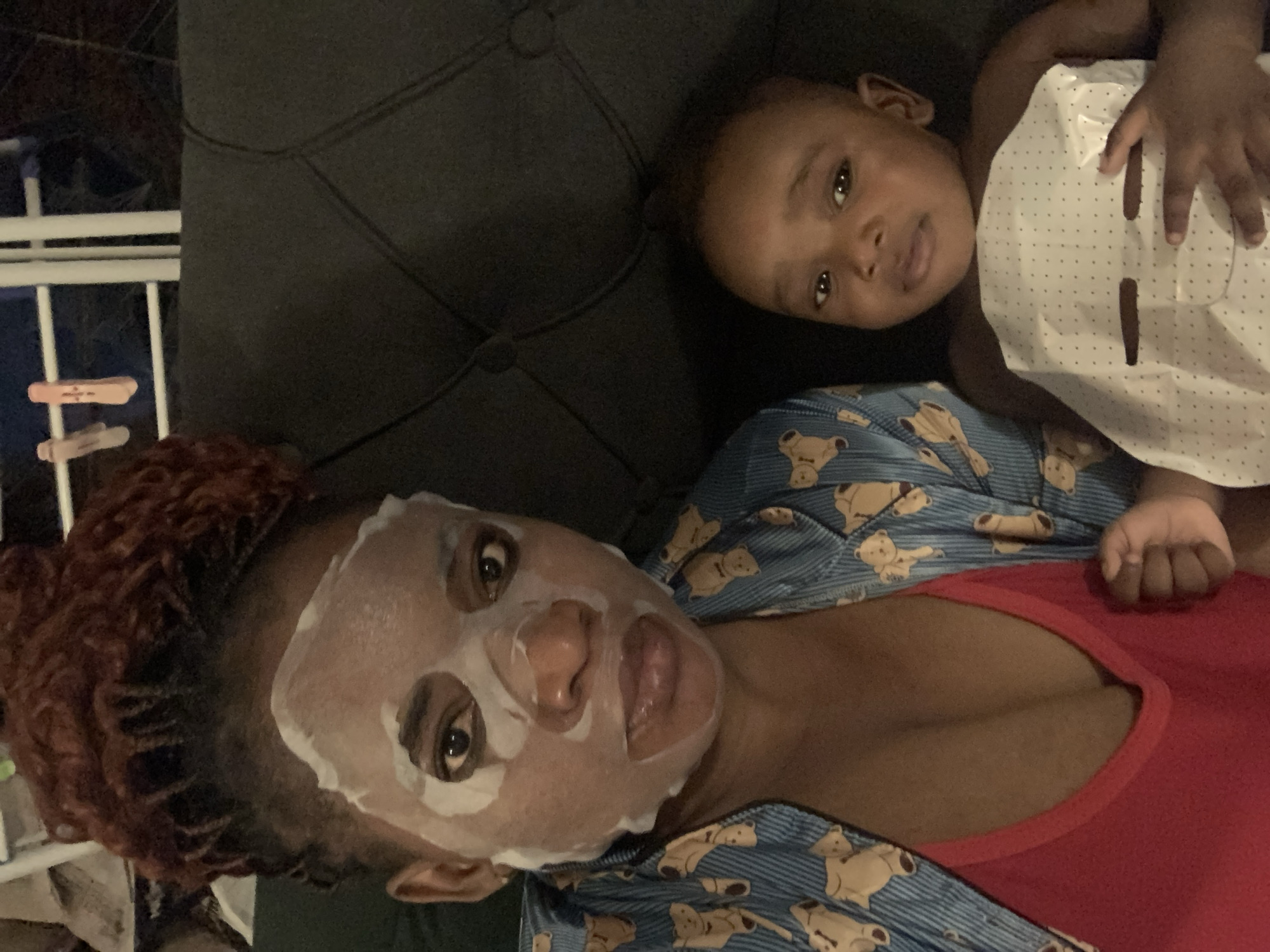 A woman and a baby are lying on a dark gray, tufted sofa. The woman has reddish-brown curly hair and a white sheet face mask on her face. She is wearing a red top and a blue pajama top with a teddy bear pattern. The baby is lying near her head and has a white face mask with black polka dots lying on their torso. The setting appears to be indoors, with a white fence-like structure visible in the background, and it is probably in a private residence. The location is identified as Barakin Akawo, Nigeria. No specific details within the image corroborate this claim.