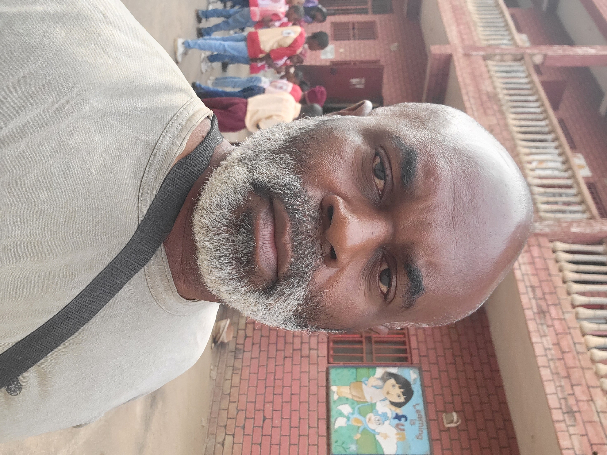 A medium close-up shot shows a man with dark skin, a bald head, and a graying beard, wearing a light-colored t-shirt with a dark strap over his shoulder. Behind him, a group of children are lined up outdoors, wearing what appear to be school uniforms. In the background, there's a building with a red brick facade and white architectural details. A colorful sign with cartoon figures is attached to the wall. The scene is located in Maiburiji, Nigeria.