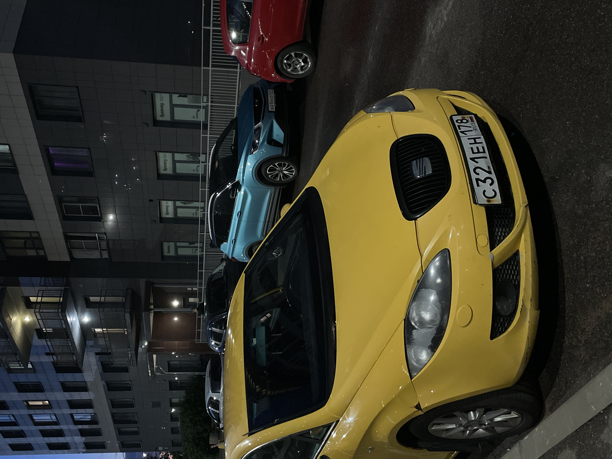 This media file captures a nighttime scene in Sinyavino, Russia, featuring a row of parked cars outside a modern building. The primary focus is a bright yellow car in the foreground, angled towards the viewer. Its headlights are on, illuminating the asphalt. Behind it, a light blue car and a red car are parked, partially visible.
The building's facade is characterized by dark grey, textured tiles and numerous windows, some of which are lit, suggesting occupancy within. Balconies with metal railings are also visible on the building's exterior. The time of day is clearly night, indicated by the darkness and artificial lighting. The weather appears to be clear.
A notable detail on the yellow car is its license plate, which reads "C321EH 178 RUS". Another license plate is partially visible on the blue car, reading "A414KO 178".
The scene depicts a quiet parking area at night, with no people visible. The composition and lighting suggest a still and perhaps somewhat solitary atmosphere. The setting is urban or suburban, indicated by the presence of a multi-story building and paved parking area.