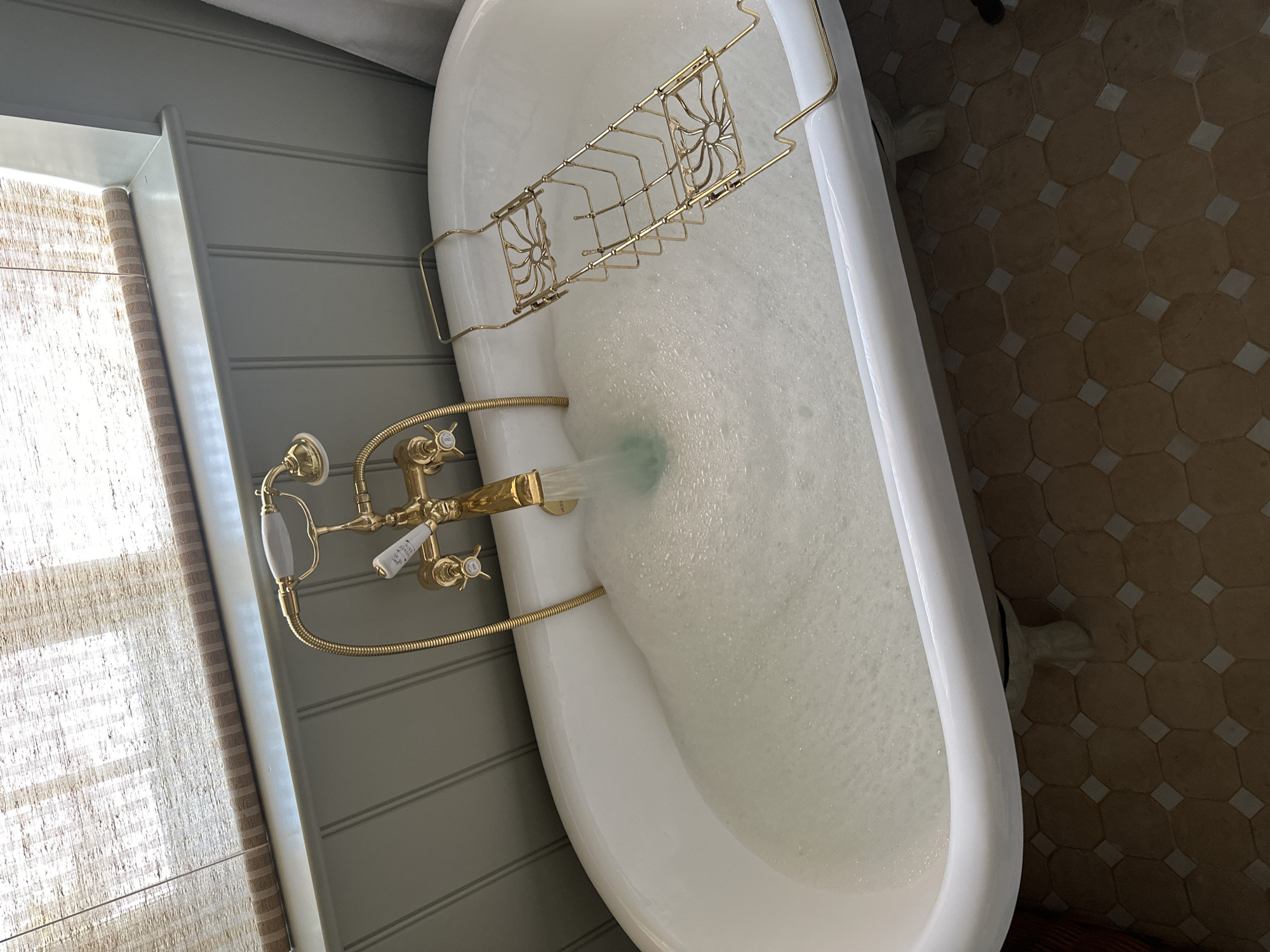 A white bathtub is nearly full of soapy water. A gold-colored bath caddy spans the width of the tub. The tub's faucet is also gold-colored and has a hand-held shower attachment. Water is running from the faucet, creating a strong stream. The wall next to the tub is gray and has horizontal paneling. A window with tan blinds is visible next to the wall. The floor is covered in tan and white hexagonal tiles. The location appears to be a bathroom in an unknown city.