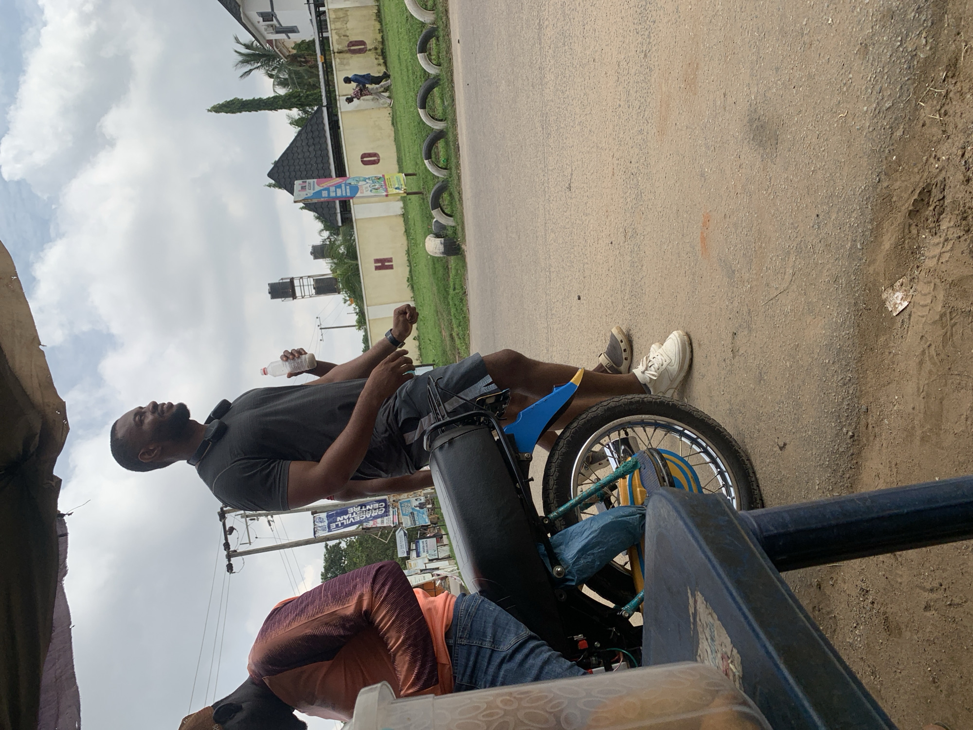 The image captures an outdoor scene under an overcast sky, likely in Abuja, Nigeria. In the foreground, a dark blue plastic table with a textured surface is partially visible on the left. Behind it, a portion of a motorcycle is prominent, featuring a black seat, a bright blue fender, and a silver-spoked wheel with yellow and blue accents. A person, partially visible, is seated on the motorcycle, wearing blue jeans and an orange and purple patterned long-sleeved shirt. To the right of the motorcycle, a man stands, wearing a dark grey t-shirt, grey shorts, and white sneakers. He holds a white plastic bottle in his right hand and appears to be looking to his right. Another person is partially visible behind him. The background shows a light-colored wall with large, red-painted letters "H" and "O" visible. Beyond the wall, there are trees, a tall building with a distinctive pyramid-shaped roof, and a water tower. A few individuals are walking in the distance near the wall. Several old tires are arranged in a line along the grassy area in front of the wall, serving as a visual border. The ground is a dusty, asphalt-like surface.