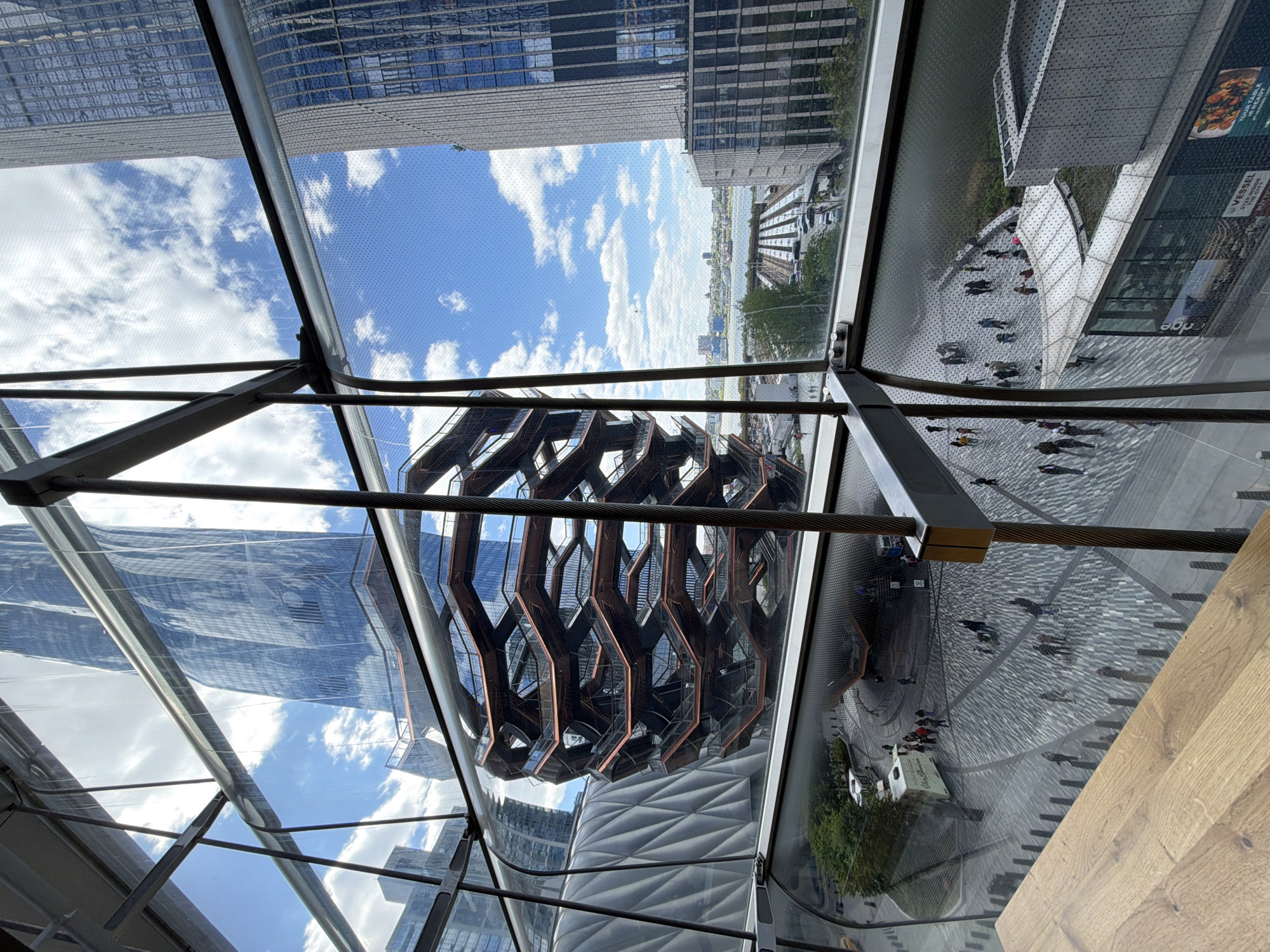 The image captures a view of The Vessel, a structure in Hudson Yards, New York City, seen through a glass or translucent roof. The roof is supported by a metal frame. Reflections of the surrounding urban landscape, including modern skyscrapers and the sky with clouds, are visible on the glass. Below The Vessel, people are walking on a patterned plaza. The Hudson River and portions of the New Jersey skyline are visible in the distance.