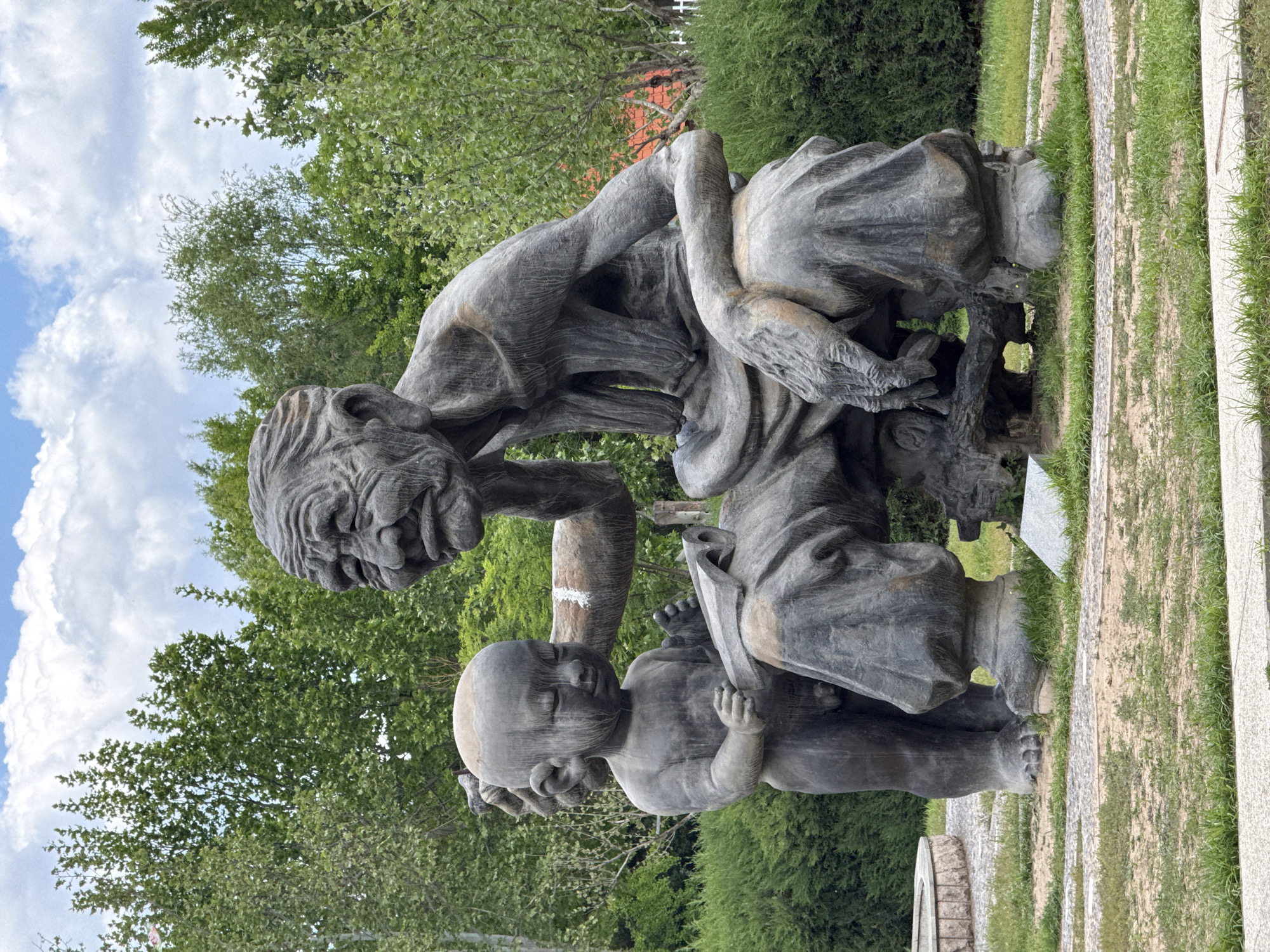 A bronze or stone statue of an old man and a child reading a book is located outside in a garden or park setting. The statue depicts an elderly man with wrinkled features and a stooped posture, looking down intently at a book held by a child sitting on his lap. The environment includes greenery, such as trees and grass, as well as some stone or concrete landscaping features. The location is in the city of Changping District, China.