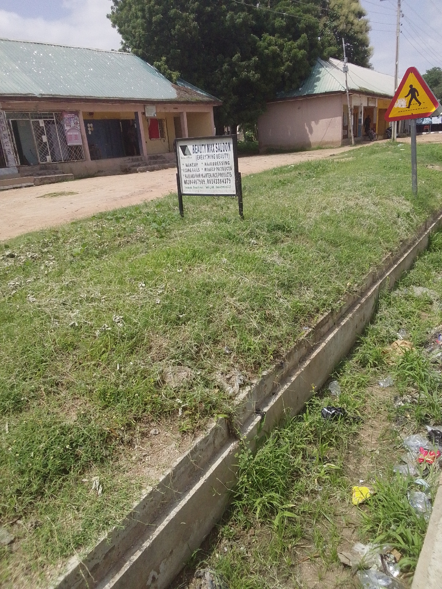 In the city of Jalingo, Nigeria, a scene unfolds with a medium shot of a street and its surrounding environment. On the left, a building with a light-colored facade and a green corrugated metal roof stretches across the frame. Its exterior features large windows and what appear to be store entrances, suggesting commercial activity. In front of this building, a dirt road is visible, with a signpost positioned to the right of center. This signpost displays a triangular yellow warning sign with a black silhouette of a pedestrian crossing.
Further to the right, another building with a similar architectural style, also featuring a green corrugated metal roof, is partially visible. In front of this second building, a single person stands near an entrance. Beside the road, a concrete drain runs parallel to the street, filled with overgrown grass and some visible debris, including plastic bags and other litter. The overall environment suggests a somewhat neglected urban area, with signs of both commerce and environmental challenges.