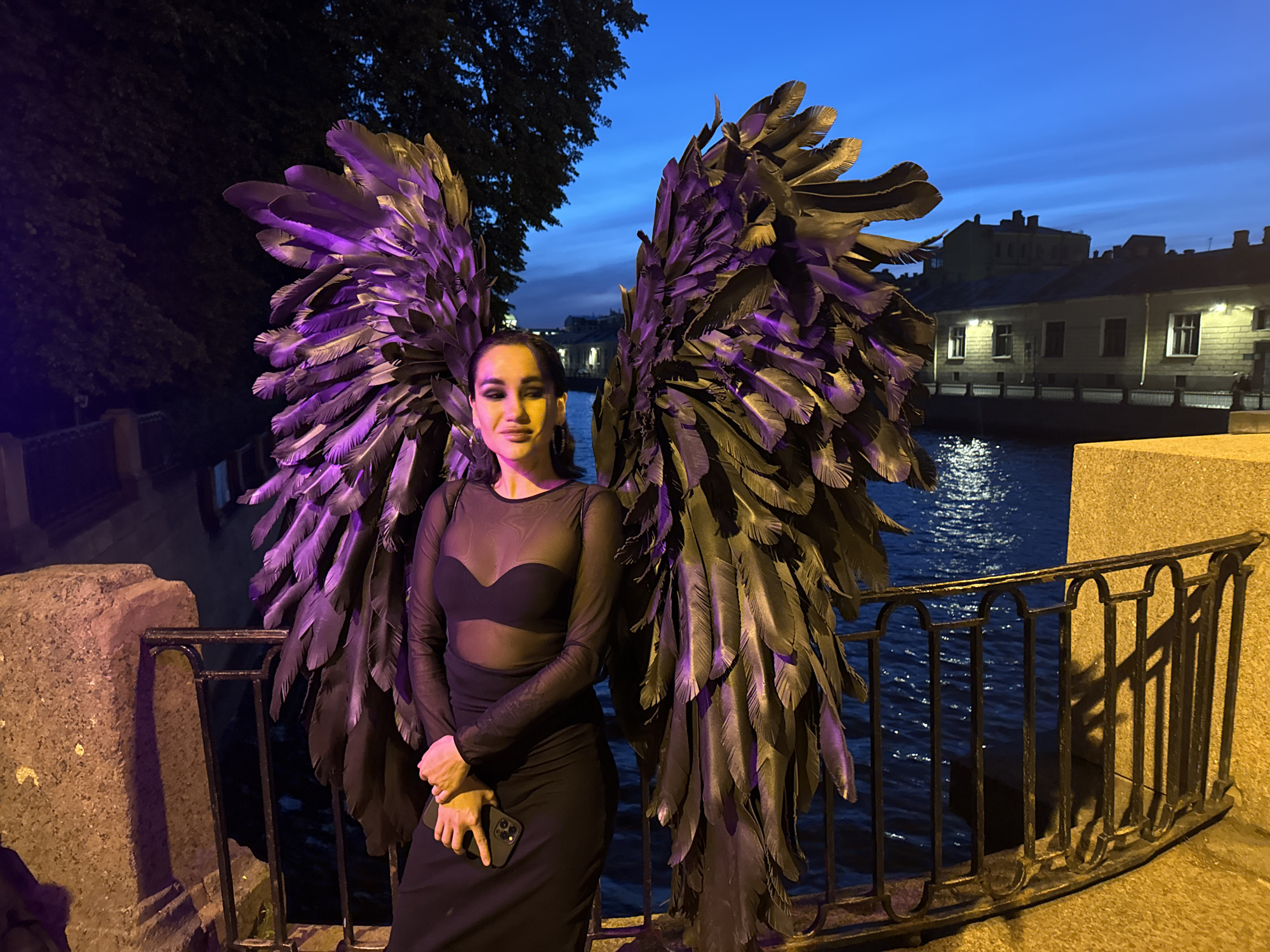 The image depicts a woman posing outdoors at night or dusk in Saint Petersburg, Russia. She stands centrally, facing forward with a slight smirk, wearing a long-sleeved black sheer top over a black bra and a long black skirt. Her hands are clasped at her waist, holding a dark-colored smartphone. Large, dark, costume wings, composed of numerous feather-like structures, extend from her back, exhibiting prominent purple illumination across many feathers, particularly on the left wing and upper sections.
The background features a deep blue twilight sky above a dark water body, likely a canal or river. Along the far bank, multi-story buildings with illuminated windows and facades are visible. To the left, dark tree foliage is present. The woman is positioned on a stone embankment or bridge, delineated by a dark metal railing with arched balusters between her and the water. A stone pillar stands to her left, and the ground surface to her right is illuminated with a yellowish light. The woman's face and hands are also lit by a yellowish light source.