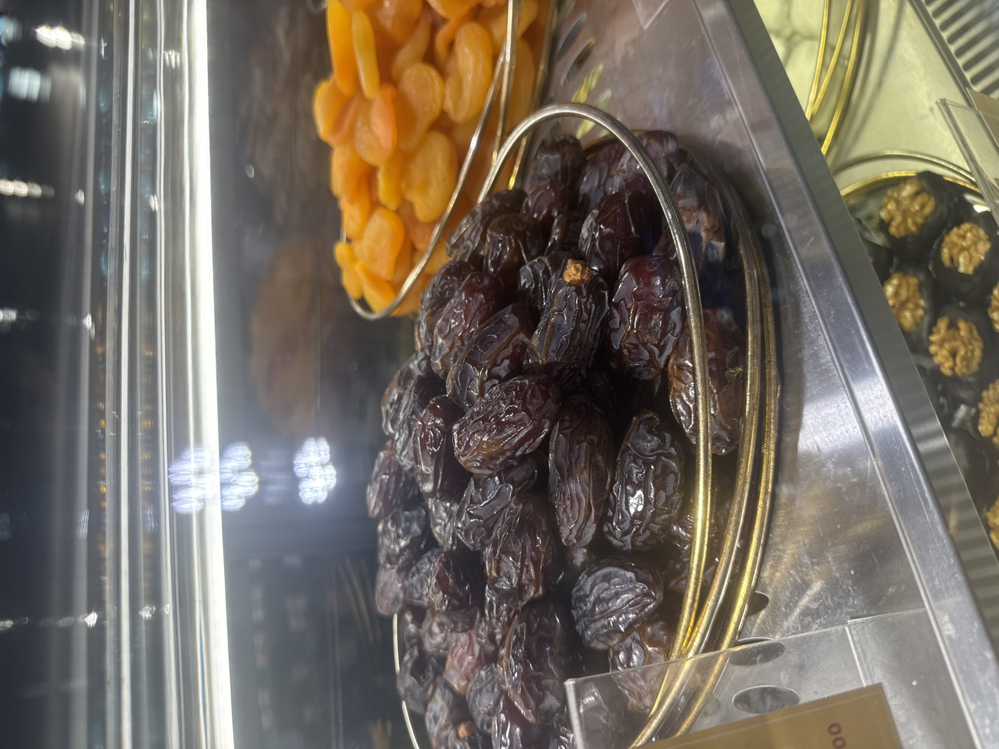 The image displays a vibrant selection of dried fruits and confections, presented in a retail display case, likely within a shop or market in Kuta Selatan, Indonesia. The primary focus is on a large quantity of dark, glossy dried dates, possibly Medjool, piled high in a gold-colored wire basket. Their wrinkled texture and rich color suggest ripeness and quality.
Behind and slightly above the dates, another gold-colored wire basket is filled with bright orange dried apricots, offering a contrasting color and texture to the scene. To the right of the dates, partially visible in the background, are dark, round confections, each topped with a golden-brown walnut half, arranged in a shallow dish, hinting at chocolate truffles or similar treats.
The items are enclosed within a clear display unit, which shows reflections and streaks of artificial indoor lighting, creating a bright and clean presentation. Metallic shelving and dividers are visible, separating the various products. The overall impression is one of neatly organized goods available for purchase. No people are visible in the frame, allowing the focus to remain entirely on the food items. In the bottom right corner, a small part of a price tag or label is visible, showing the numbers "00" oriented vertically. The image is taken from a slightly elevated perspective, looking down into the display.