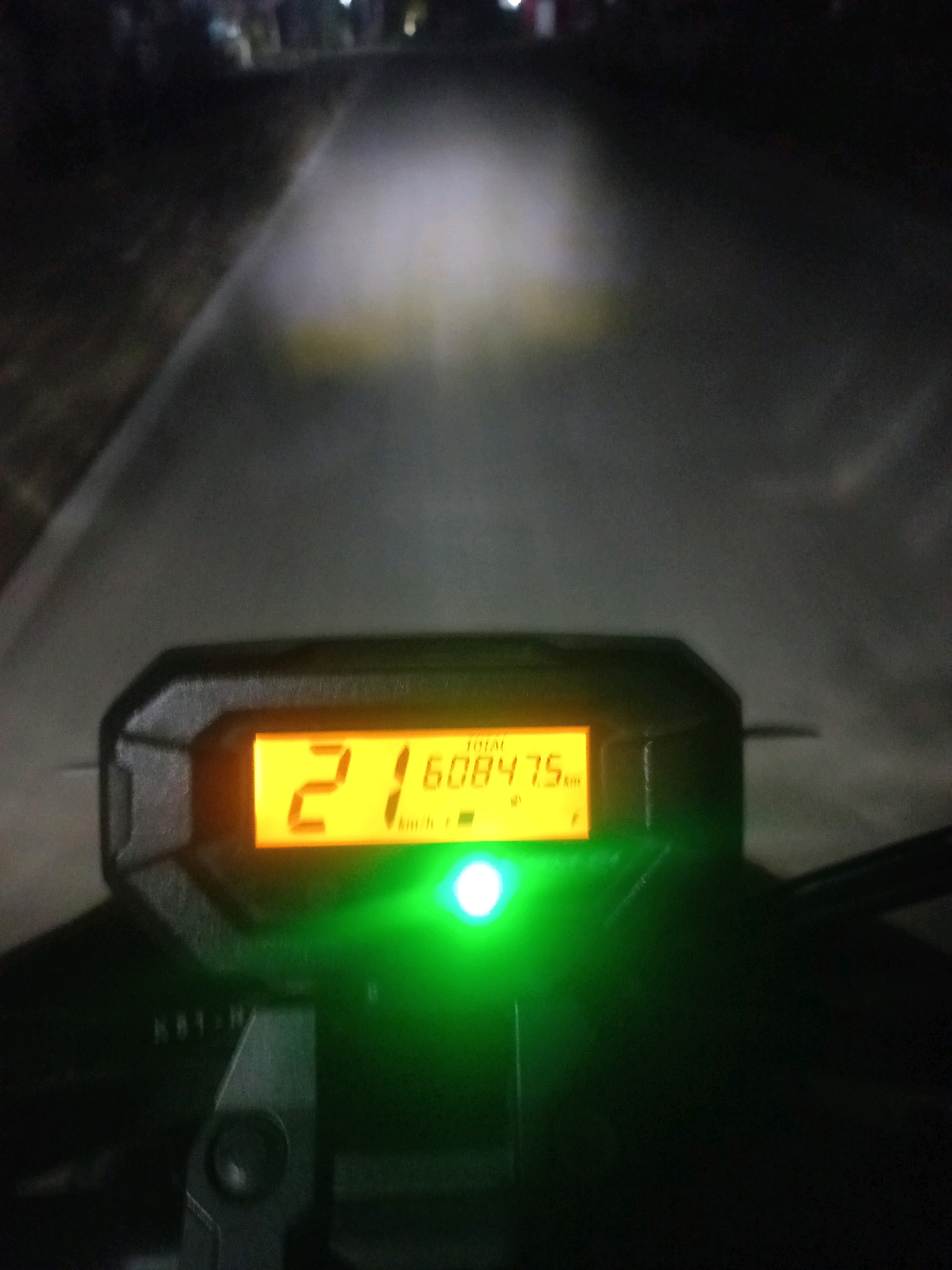 The image shows a close-up view of a motorcycle's digital dashboard at night. The dashboard displays the word "TOTAL" above a numerical reading of "21 60847.5" followed by "km/h". A green indicator light is illuminated below the display. The background is blurred, suggesting motion, and depicts a dimly lit road or path with some ambient lights visible in the distance, consistent with an outdoor, possibly rural, environment. The location is identified as Gambiranom, Indonesia, based on the provided context.