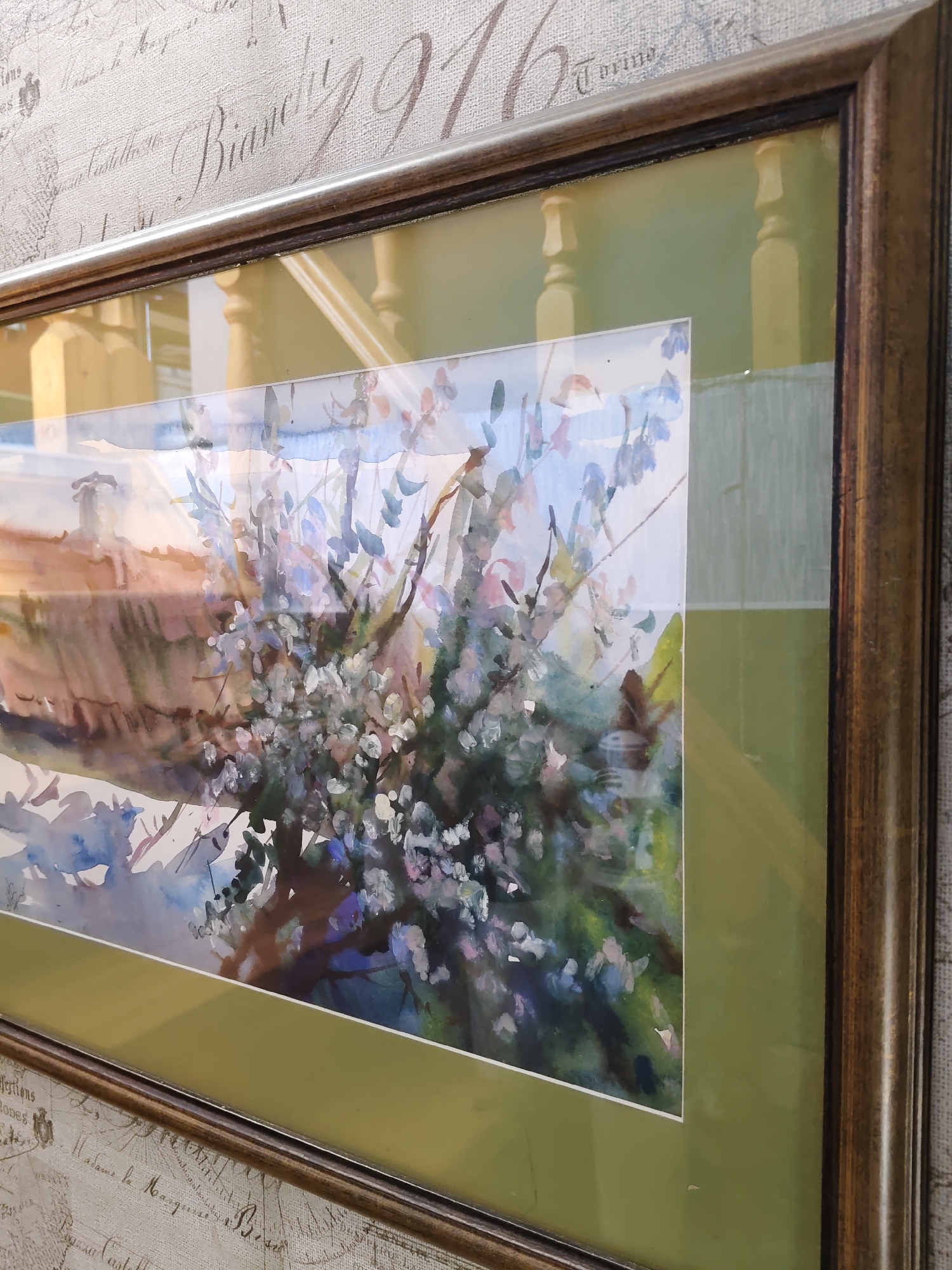 The image shows a framed watercolor painting of a tree with light pink and white blossoms against a backdrop of a building with a red roof and a chimney. The painting is framed in a dark wooden frame with a lighter colored matting. The background of the photograph shows a textured wallpaper with faint script writing and a wooden railing. The photograph is taken at a slight angle, with the bottom left corner closer to the viewer than the top right. The lighting suggests an indoor environment. The provided crops show details of the frame, the wallpaper, and sections of the painting. There is no information or visual element within the image that indicates the location is the city of Rostov-on-Don, Russia.