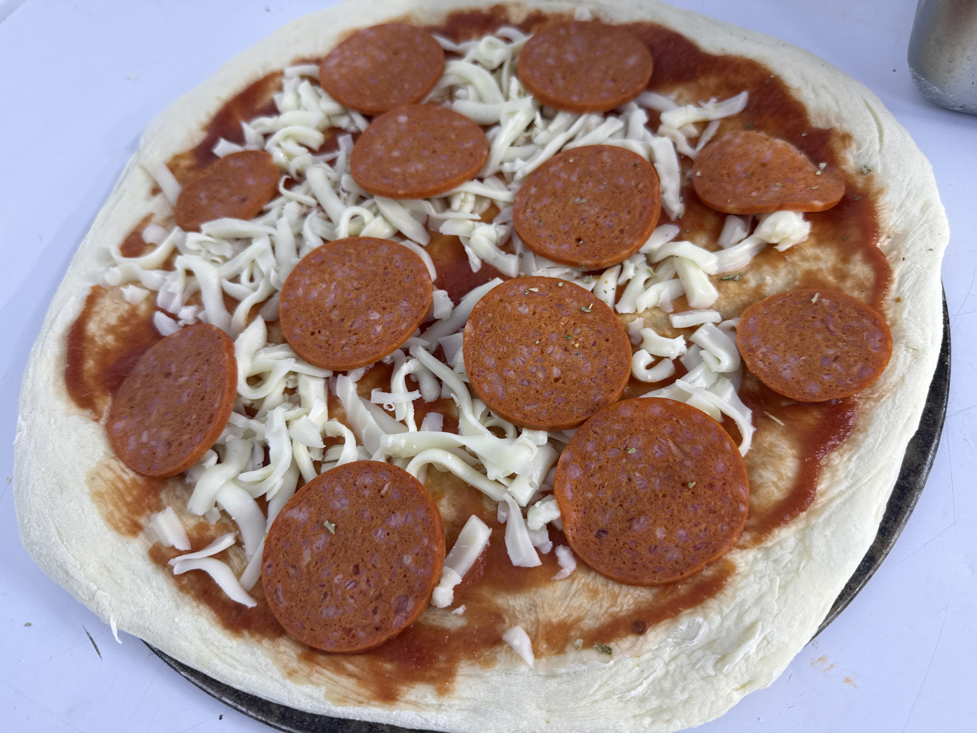 The image shows an uncooked pizza with pepperoni and shredded mozzarella cheese on a white surface. The pizza is round and uncooked, with a thick crust. The sauce is spread evenly across the dough, and the pepperoni slices are arranged in a circular pattern on top of the cheese. The mozzarella cheese is shredded and appears to be fresh. The pizza is on a dark-colored pizza pan, which is partially visible at the bottom of the image. The background is a light-colored, possibly white or light gray surface, with some blurry elements that could be part of a kitchen or outdoor setting. There are no people visible in the image. The scene is static, depicting the preparation of a pizza. The location in Akshi, Kazakhstan, is not evident from the visual information in the image.