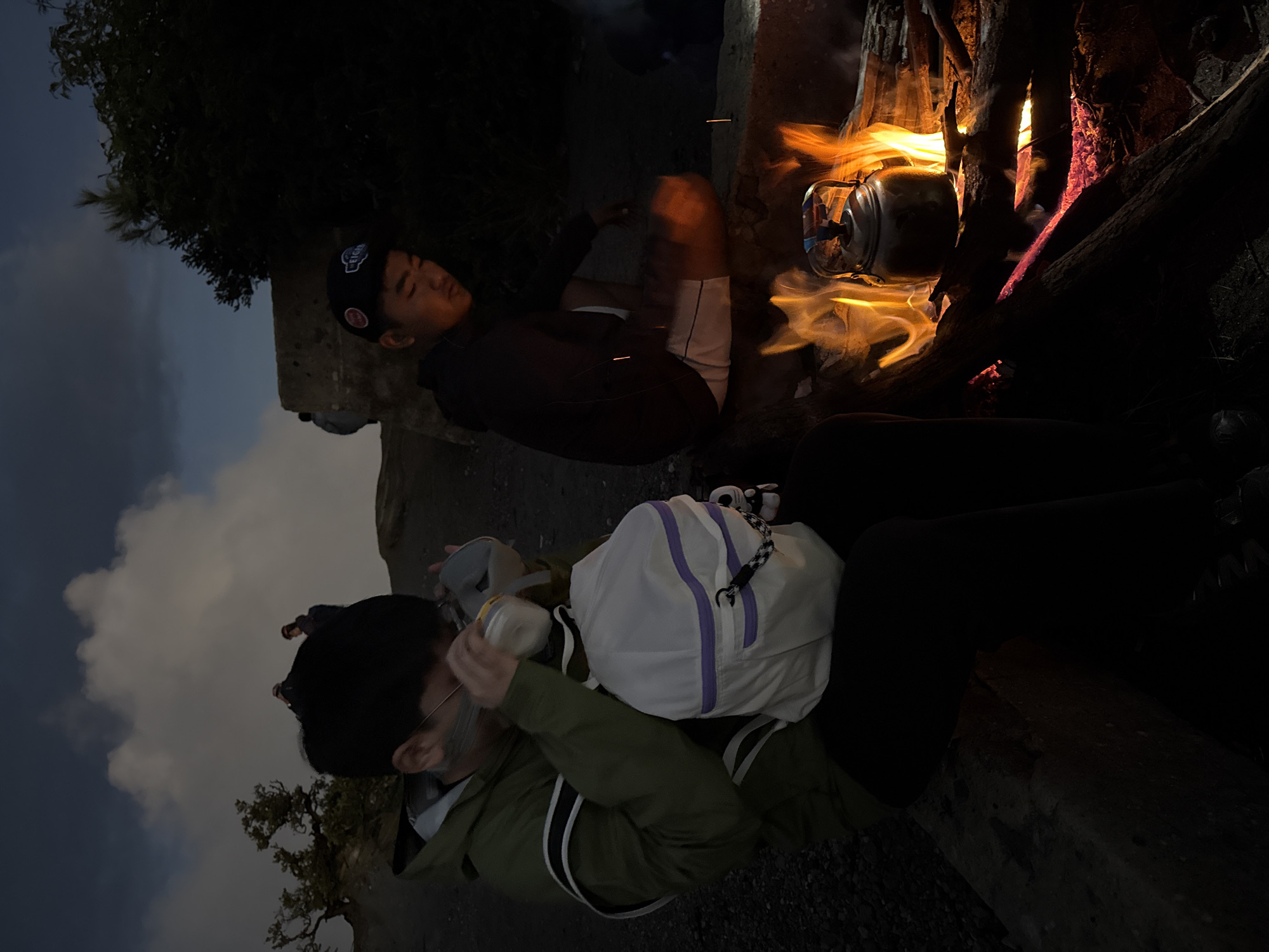 The image shows two people near a campfire at night, possibly outdoors in a rugged area. The person closer to the viewer is wearing a green jacket and holding a bottle, perhaps drinking. The other person is lying down near a tree and the fire, with their legs bent. A silver kettle sits in the fire. The environment includes trees and what appears to be rock, suggesting a natural setting. It's dark, with light provided by the fire.