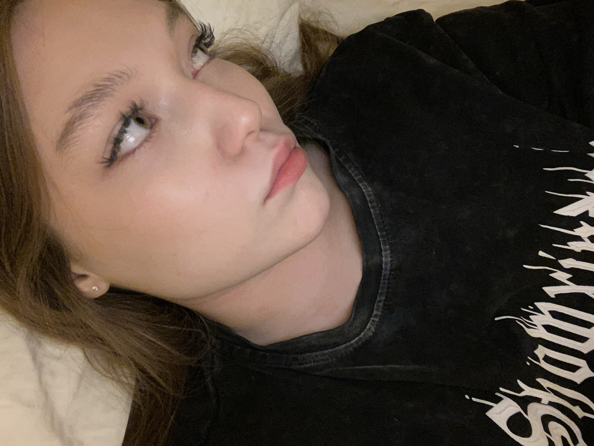 A young, light-skinned woman is lying down, presumably on a bed, with her head resting on a white pillow or bedding. She is wearing a dark-colored t-shirt with a white graphic design visible on the chest area. The woman is angled towards the viewer, her gaze directed upwards and to the left. Her hair is light brown, and her facial features are clearly visible, including her eyes with enhanced eyelashes, nose, and lips. A small earring is visible in her left ear. The lighting is soft and diffuse, suggesting an indoor setting. There is no indication of location in Saint Petersburg, Russia.