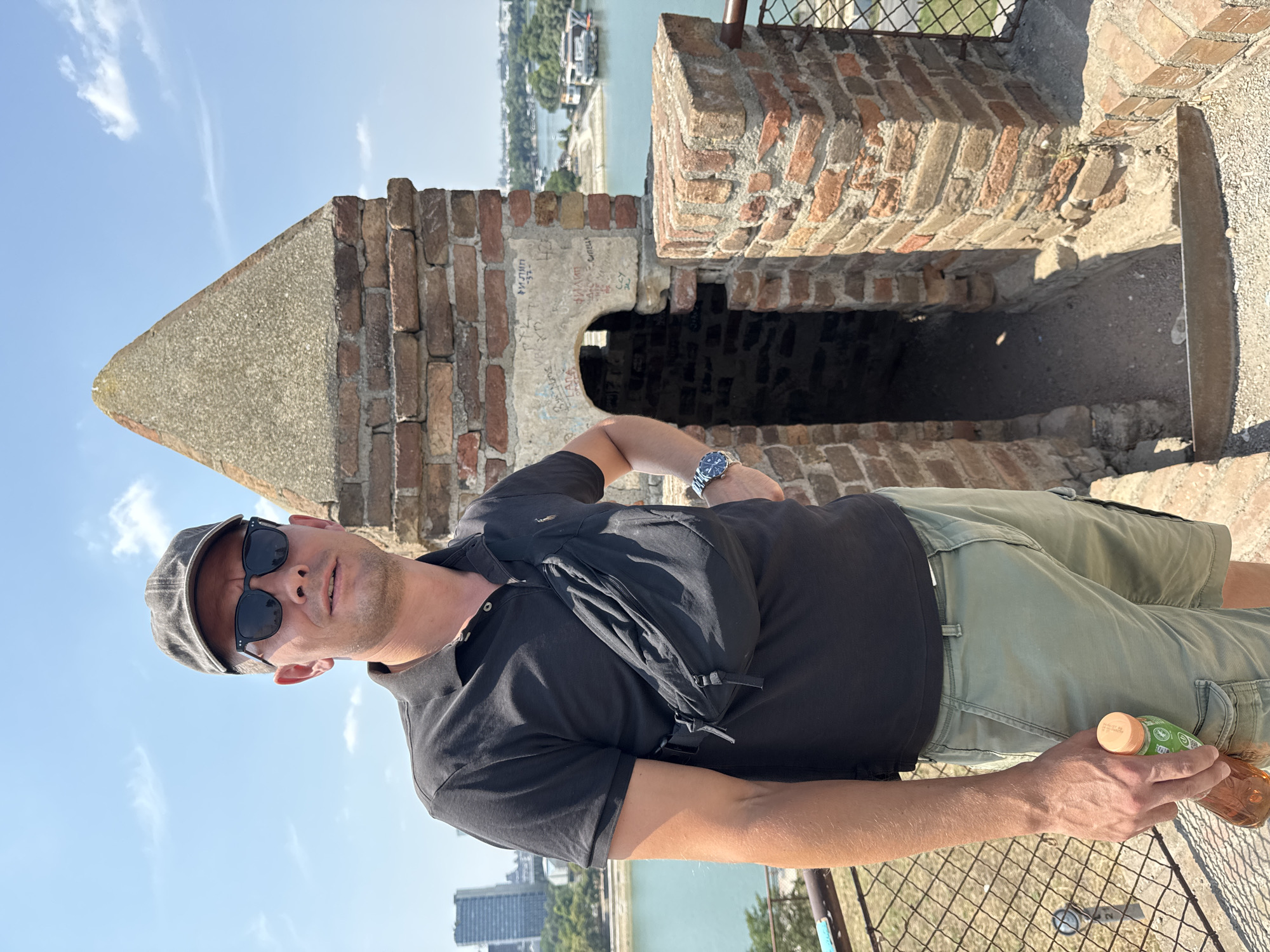 A man stands on a rampart at the Belgrade Fortress, Kalemegdan, Belgrade, Serbia. He is wearing a dark polo shirt, khaki cargo shorts, a gray baseball cap, and dark sunglasses. A black fanny pack is worn across his chest. He is holding a bottle of amber liquid with a green and white label in his right hand. He is looking towards the camera with a neutral expression. Behind him, to the right, is a section of the fortress wall made of red brick with an arched opening. Beyond the fortress walls, a body of water, likely the confluence of the Sava and Danube rivers, is visible, with boats and buildings on the far bank. The sky is clear and blue with scattered clouds. The foreground shows a metal railing and a grated fence.