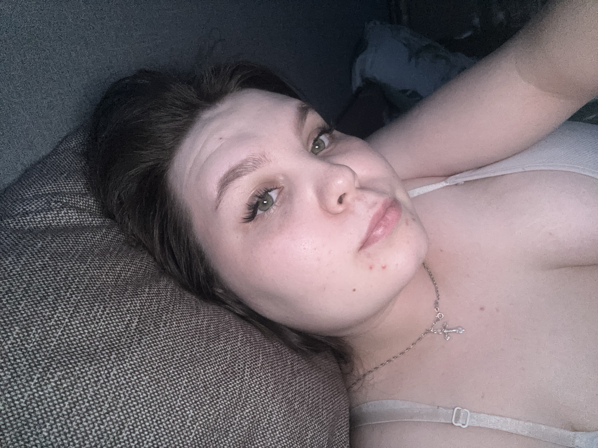 The image captures a close-up, slightly angled portrait of a person, likely a woman, lying on her side. Her head is resting on a textured, dark grey or brown fabric, which appears to be a couch or a large cushion.
The person has a fair complexion with visible freckles and a few small red blemishes on her chin/jawline. Her eyes are a striking green, and her eyebrows are dark and well-defined, possibly filled in. She has long, dark eyelashes, which appear to be extensions, framing her eyes. Her dark hair is styled away from her face. Her lips are slightly parted, showing a hint of a light pink or nude lip color, and she has a relaxed expression, looking directly at the camera with a subtle hint of a smile or smirk. Faint lines are visible on her forehead.
She is wearing a light-colored (possibly white or light beige) bra, with a visible strap and metal clasp. A delicate silver-colored necklace with a small cross pendant hangs around her neck, resting on her upper chest. Her right arm is visible, bent and extending upwards or behind her head. The lighting in the image is somewhat dim and soft, creating a calm and intimate atmosphere, suggesting an indoor setting. The fabric of the couch/cushion has a distinct woven, speckled pattern. No text is visible in the image.