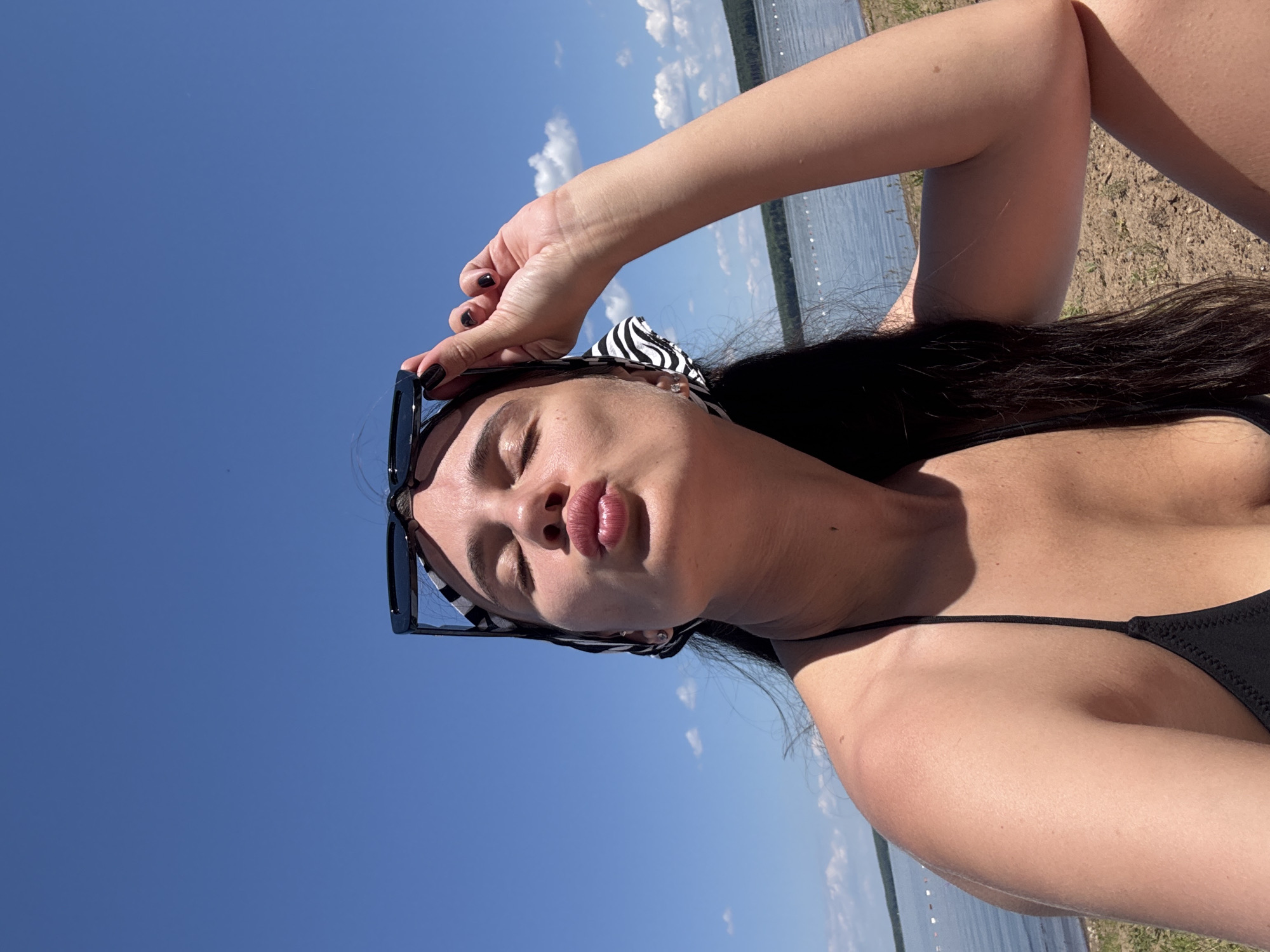 In a sunny, outdoor, daytime scene, a young woman with long, dark hair, wearing a black bikini top and a black and white striped headscarf, sits on a sandy beach. She is holding her sunglasses above her head with her right hand, her eyes are closed, and her lips are puckered in a kissing gesture. Her left arm is bent at the elbow, resting on her lap. Behind her, a body of water stretches to the horizon, with a tree-lined shore visible in the distance. The sky is a clear, bright blue with a few scattered white clouds. The location appears to be the Kama River embankment in the city of Chaikovsky, Russia, given the characteristic landscape of this area.