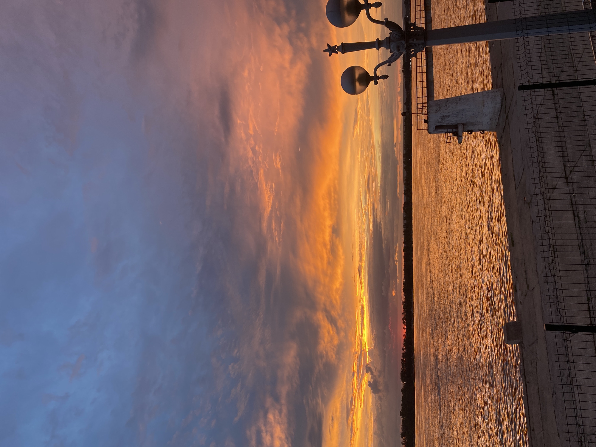 The image depicts a scenic sunset over a wide river, likely the Volga River, near Novokuybyshevsk, Russia. The sky is a vibrant display of orange, yellow, and pink hues, with scattered clouds adding texture and depth. The river reflects the warm colors of the sunset, creating a shimmering, golden surface. Along the riverbank, a concrete embankment is visible, with a metal fence in the foreground. A lamppost with two ornate lamps stands on the embankment, adding a touch of urban detail to the natural beauty of the scene. In the distance, a dark line of trees marks the opposite bank of the river, silhouetted against the colorful sky. The overall atmosphere is tranquil and serene, capturing the beauty of a summer evening in Russia.