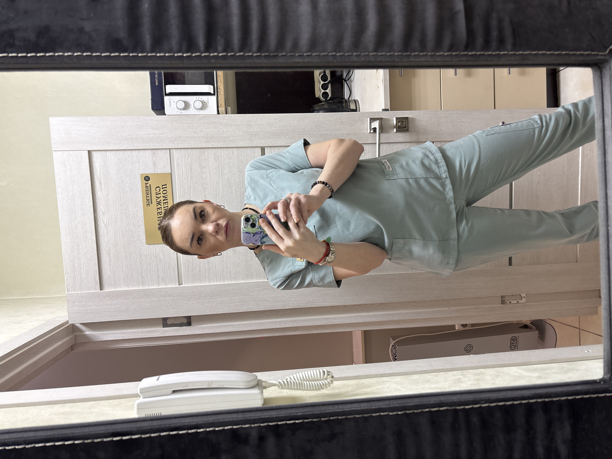 A mirror reflection reveals a woman in light blue scrubs holding a cell phone, seemingly taking a selfie. Behind her is a light-colored door with Russian text. Above the door, there is a microwave and an electrical outlet strip. Below the door, a white telephone sits on a surface, and a machine of unknown type and use is visible. The location is not identifiable.
