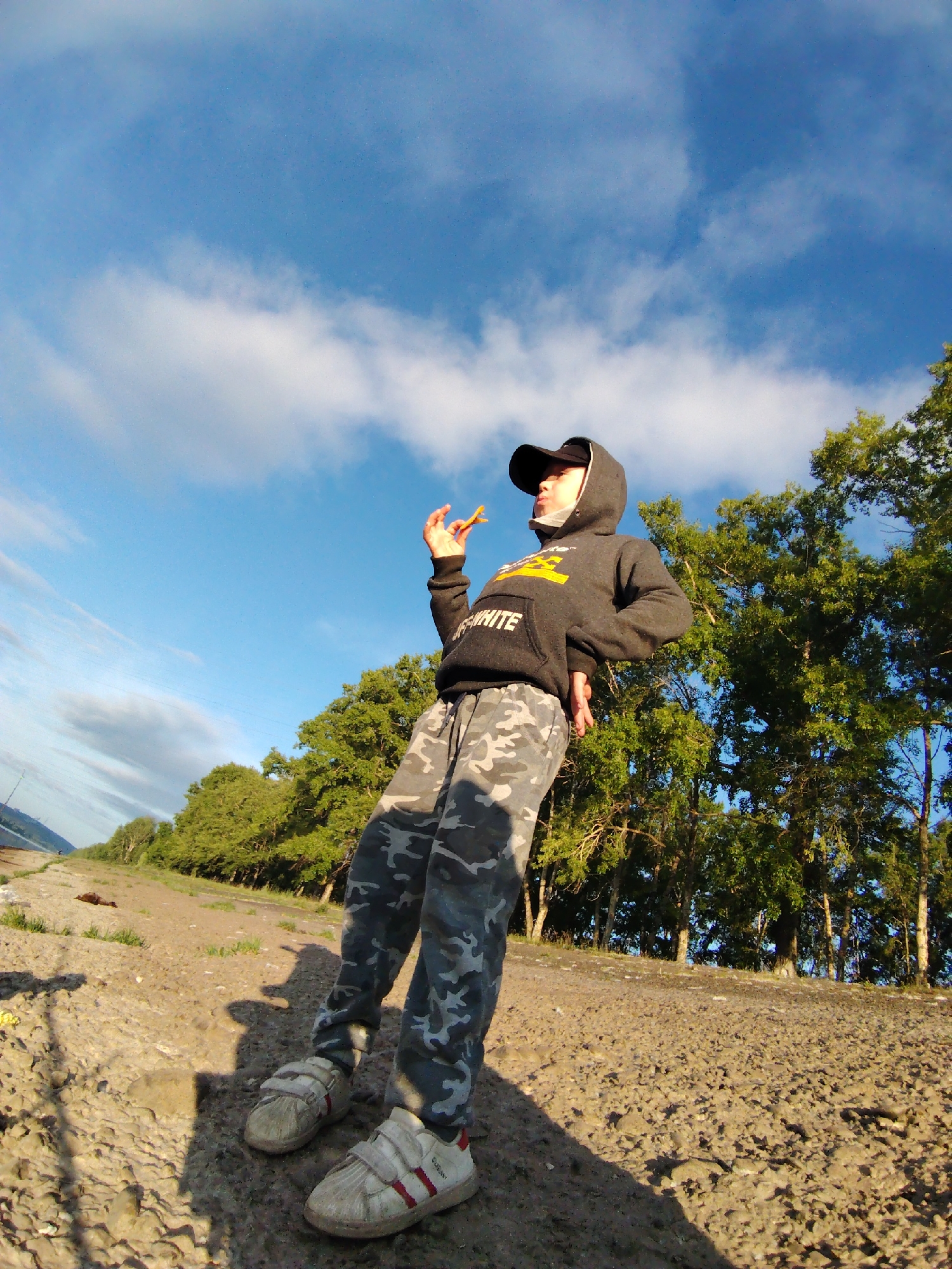 A young person, wearing a black hooded sweatshirt with yellow lettering, camouflage pants, and a black baseball cap, stands on a dirt path outdoors. They are holding a piece of orange food up to their mouth. The background features a line of green trees against a bright blue sky with scattered white clouds. The ground is covered in gravel and dirt. The image appears to be taken with a wide-angle lens, creating a slightly distorted perspective. The setting is likely a park or natural area within the city of Chaikovsky, Russia, given the vegetation and open sky.