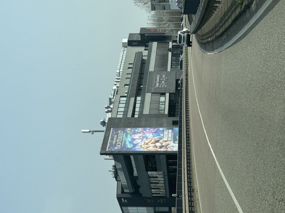 The image displays a modern, multi-story commercial building situated adjacent to a multi-lane asphalt road, in the vicinity of Altenheim, Germany. The building features a dark grey or black facade with numerous windows and recessed sections, suggesting balconies or additional architectural elements. A large vertical banner on the left side of the building advertises "Nordisches Lichterfest" and "Wasserwelt des Europa-Park," depicting individuals in swimwear alongside illuminated scenery. To the right, text signage on the building reads "Theater Eurodistrict BAden ALsace." The rooftop contains various mechanical installations, including ventilation units and communication antennas. In the foreground, a two-lane road with a solid white center line curves to the right, bordered by a metal guardrail. A white passenger vehicle is observed traveling in the right lane, moving away from the viewer. Several other vehicles are visible parked or in the distance behind the guardrail. The sky is clear and light blue, with some bare trees visible in the far background to the right.