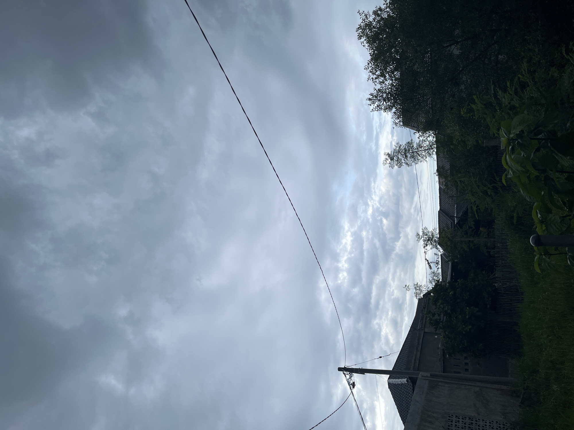 The image captures an overcast, daytime scene primarily composed of a heavily clouded sky and elements of a residential area. The sky, dominating the upper portion, is filled with dense, grey stratus clouds, suggesting either an approaching storm or simply a gloomy day with diffused sunlight peeking through in certain areas. A thin, dark power line stretches diagonally across the sky from left to right, running from a utility pole visible towards the bottom right.
In the midground and foreground, a cluster of residential buildings, likely houses, is visible. These structures appear to be made of concrete with dark tiled roofs. Dense, lush green foliage and trees partially obscure the buildings and line the right side of the frame, contributing to a somewhat overgrown appearance. A patch of green grass is visible along the very bottom right. A sturdy utility pole, with power lines extending from it, stands in the bottom right corner. The overall lighting is subdued due to the thick cloud cover, casting a soft, even light over the scene. No people or specific activities are depicted, giving the impression of a quiet, everyday street scene. There is no visible text. The architecture and vegetation align with the provided location context of Lhokseumawe, Indonesia.