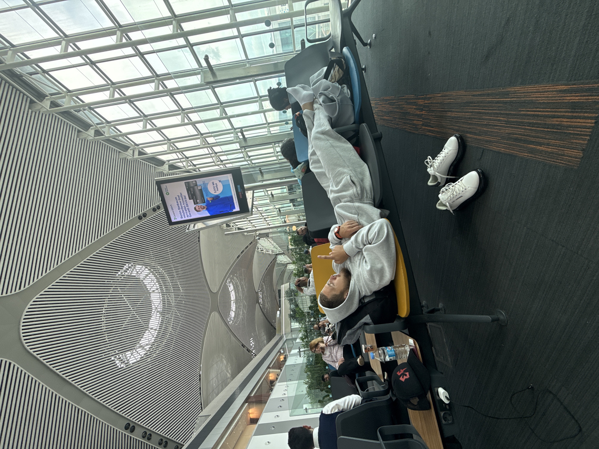 The image shows a person lying across multiple airport seats, seemingly sleeping or resting. The person is wearing a grey hoodie and sweatpants. Their white shoes are on the floor next to the seating area. A television screen displays an advertisement in Turkish. The airport has a high, modern ceiling with patterned architectural details. Other people are visible in the background, seated or walking. The setting is likely a waiting area in an airport terminal, possibly in Arnavutköy, Turkey, given the ad shown on the screen. A water bottle and a black baseball cap are next to the seating area.
