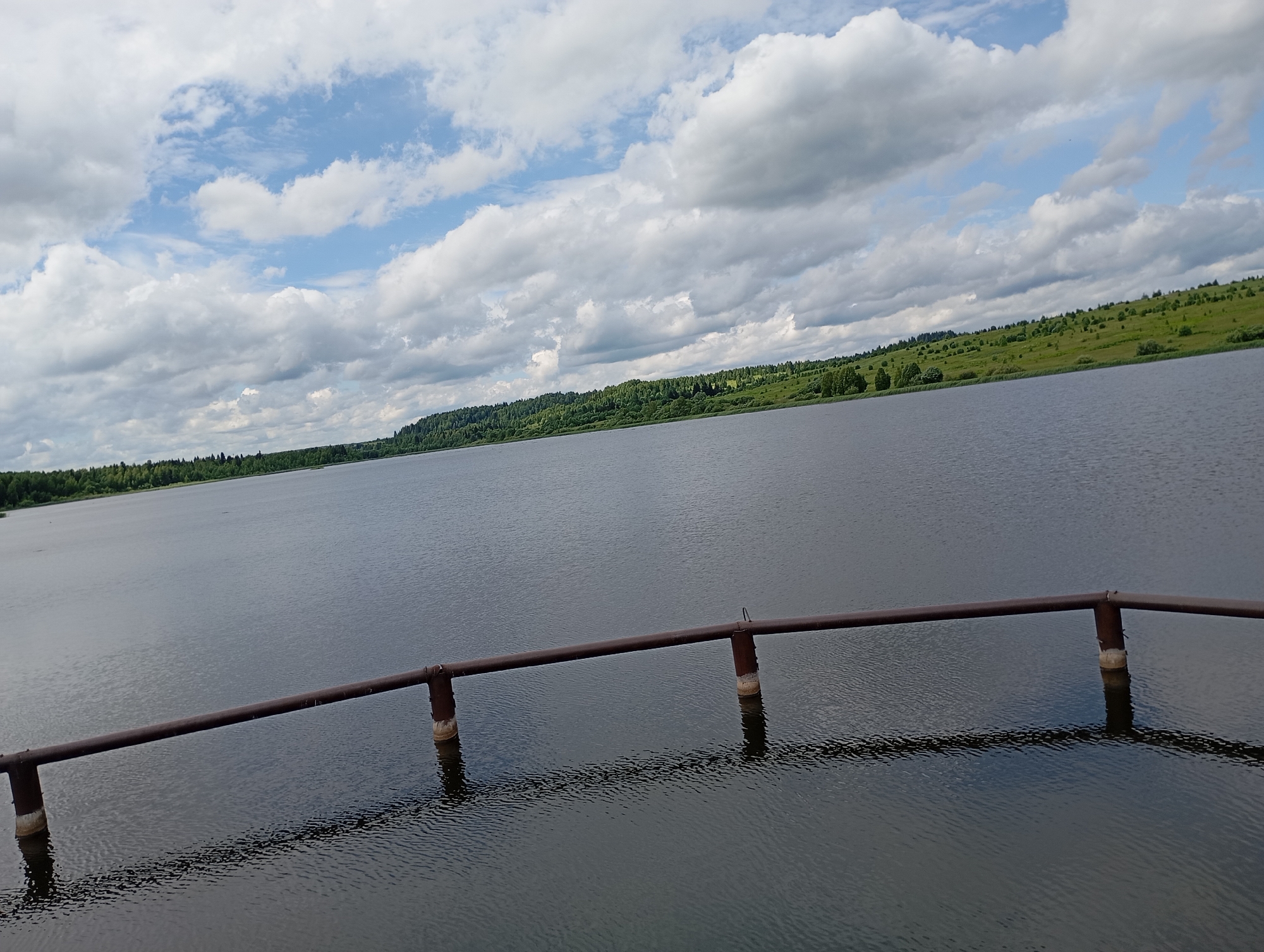This is a wide-angle photograph of a body of water, likely a lake or reservoir, with a shoreline covered in trees and green hills in the distance. The sky is filled with fluffy white clouds against a blue background. In the foreground, a rusty metal railing supported by concrete pillars extends over the water. The railing casts a reflection on the rippling surface of the water. The scene appears to be located in the village of Ryabki, Russia, given the provided context. There are no people or visible signs of activity in the image. The overall impression is one of a calm, natural landscape.